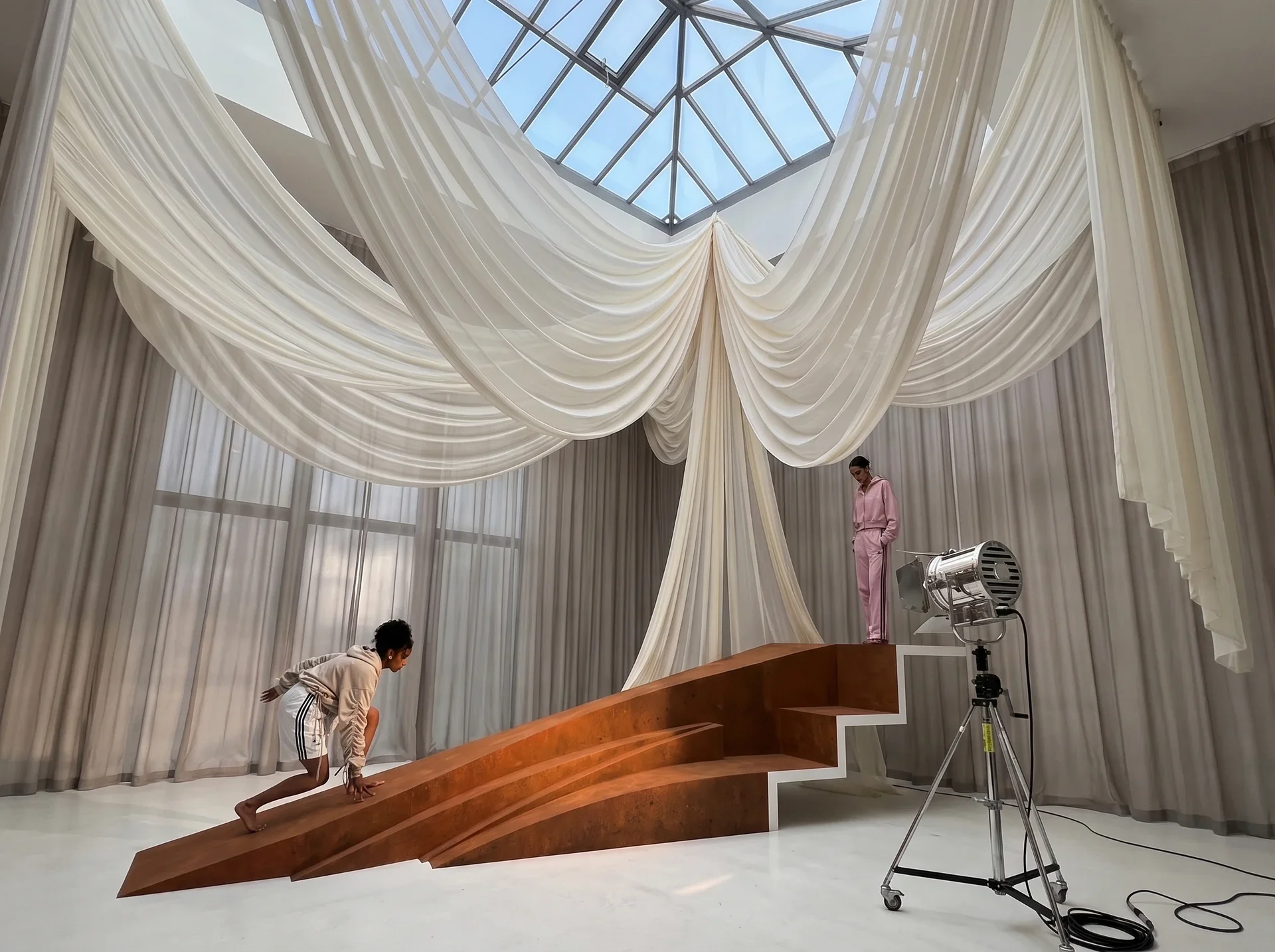 Two people, one stretching and another standing on a stage, in a room decorated with large, flowing white curtains and a glass ceiling. There is also a professional studio light in the room.