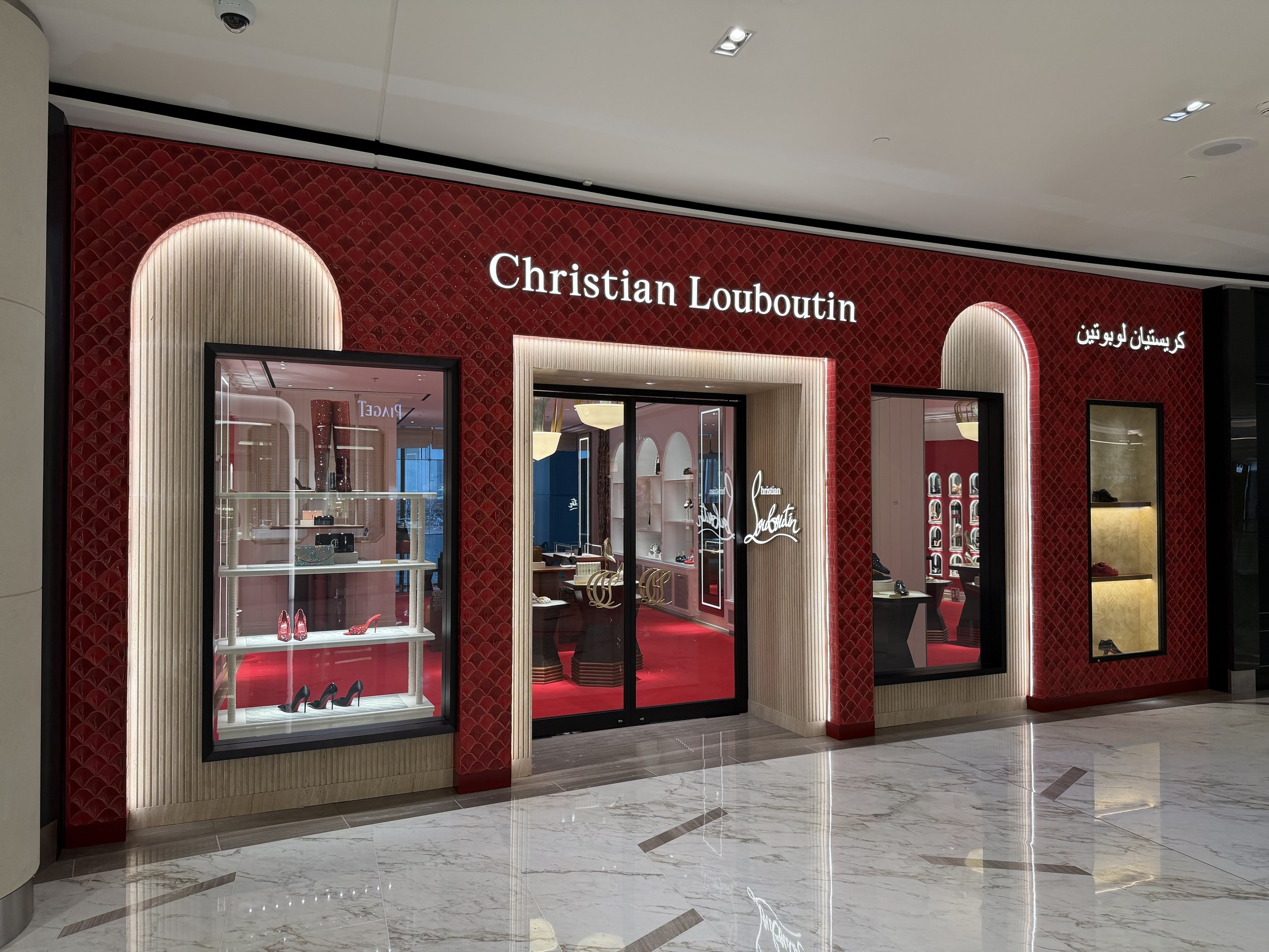 Front of Christian Louboutin store with red patterned walls and illuminated sign, display windows with shoes, marble floor.