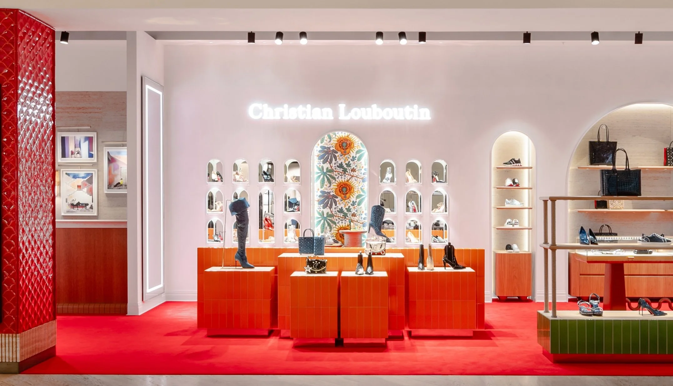 Inside a Christian Louboutin store with bright display window, shoes on orange podiums, a floral mural, and shelves with handbags and shoes.