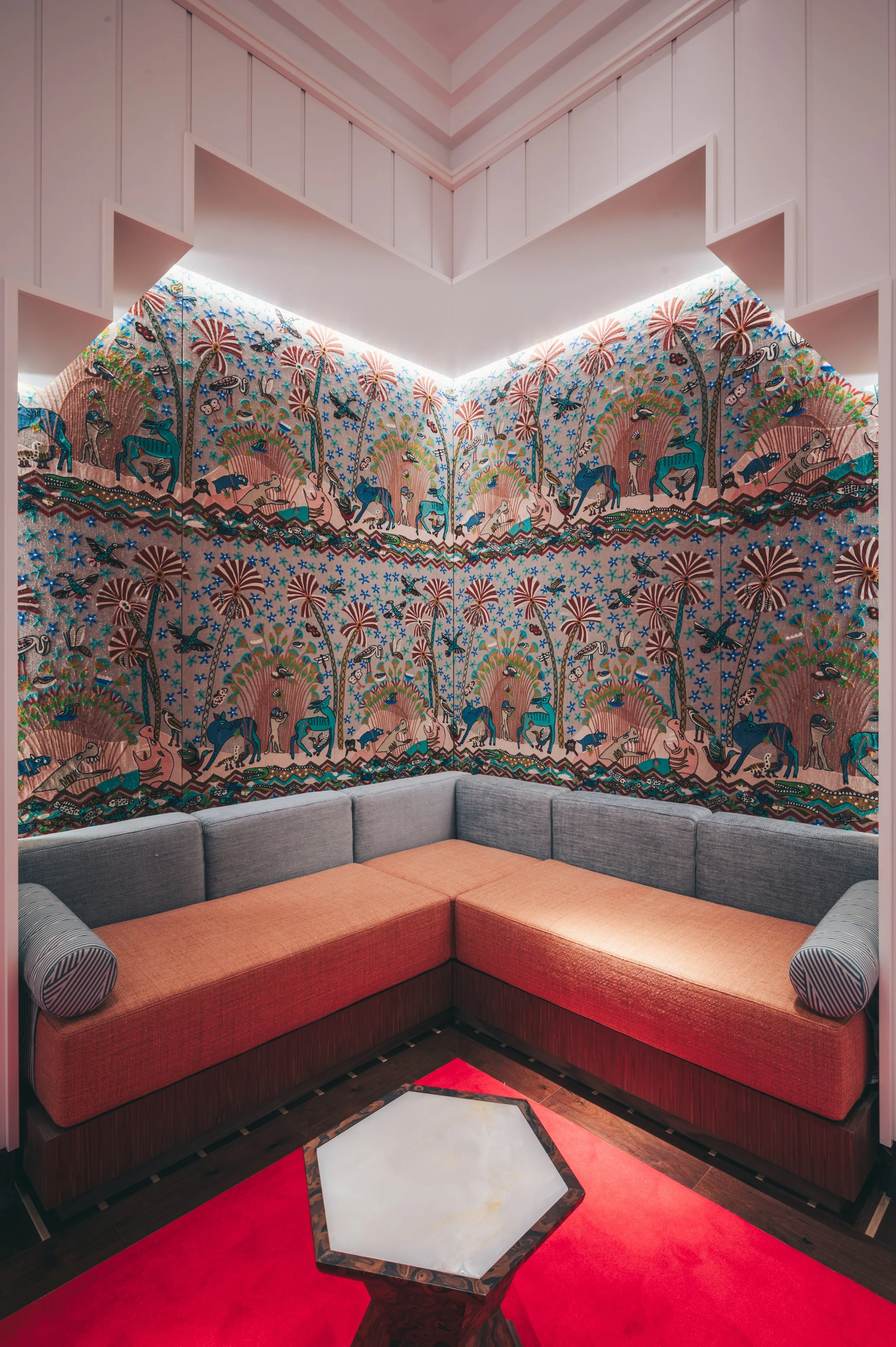 Corner of a room with a multicolored wall mural depicting animals, trees, and birds, above a gray and orange sectional sofa, with a hexagonal marble-topped side table on a red carpet.