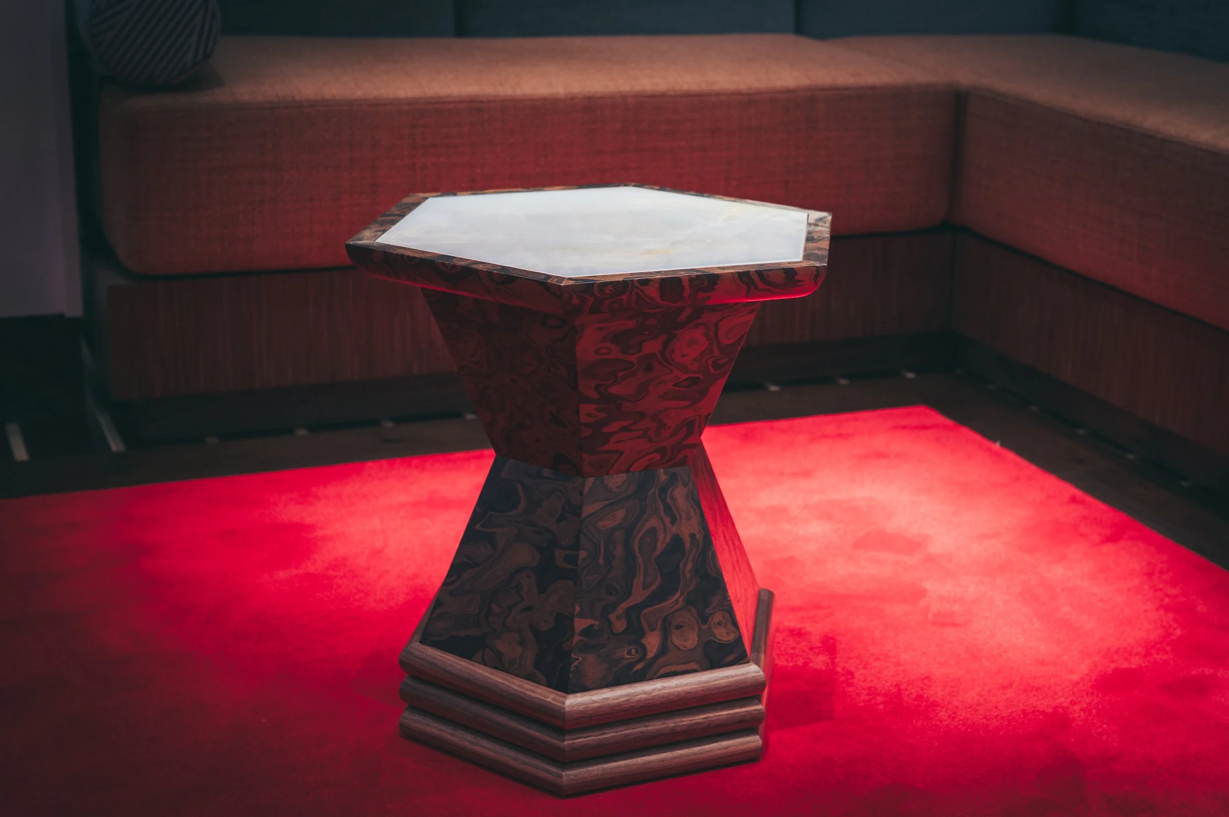 A hexagonal marble table with a wooden base in a dimly lit room with red carpet and red upholstered seating.