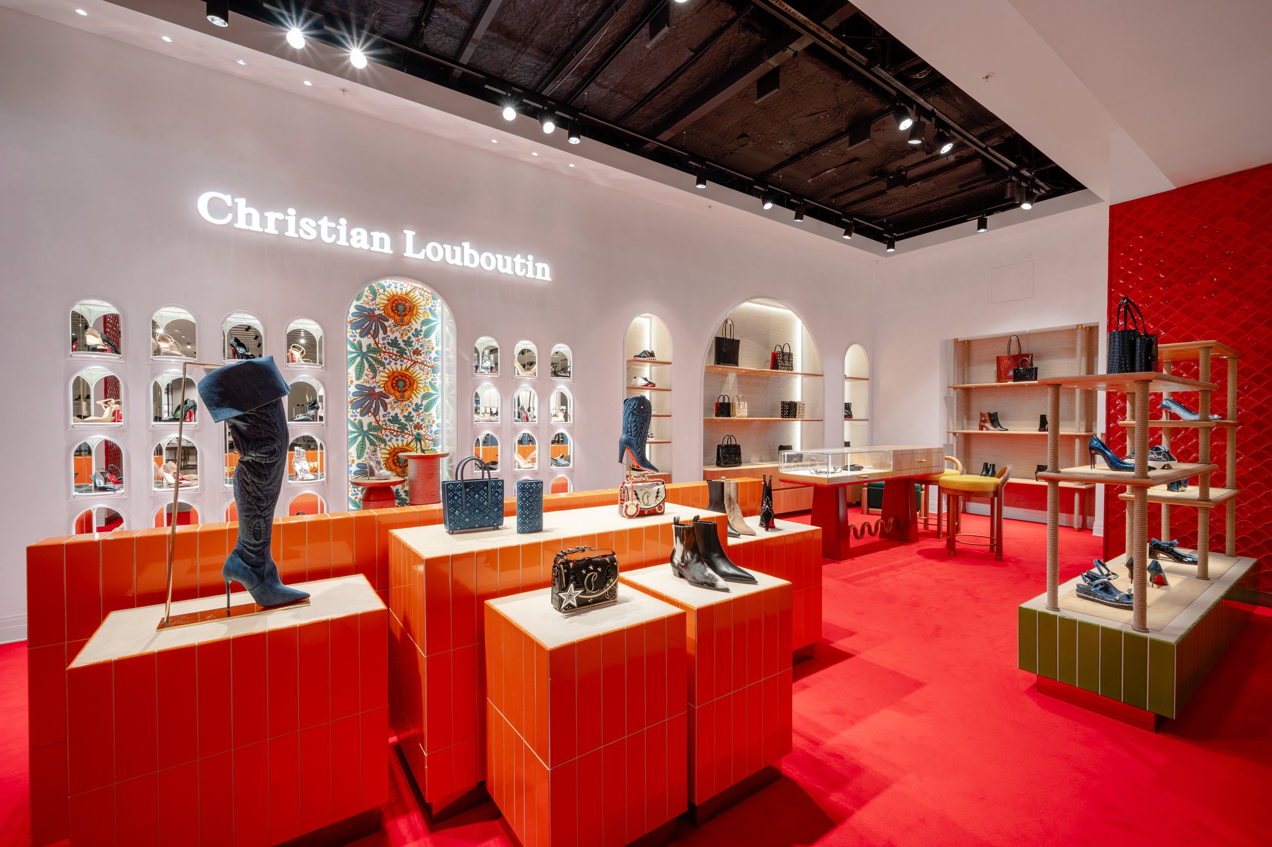 A Christian Louboutin store interior with red tiled displays, shelves with handbags, and shoes on illuminated racks, featuring a decorative floral mural, a red carpet floor, and white walls.