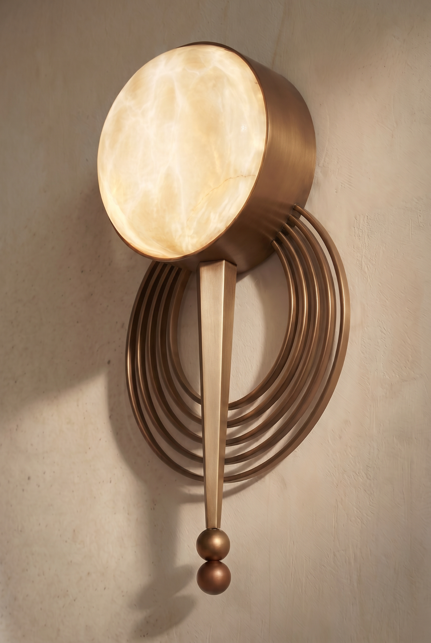 A decorative wall-mounted light fixture with a circular, glowing alabaster shade, bronze concentric rings, and three bronze spheres hanging from a bronze finial.
