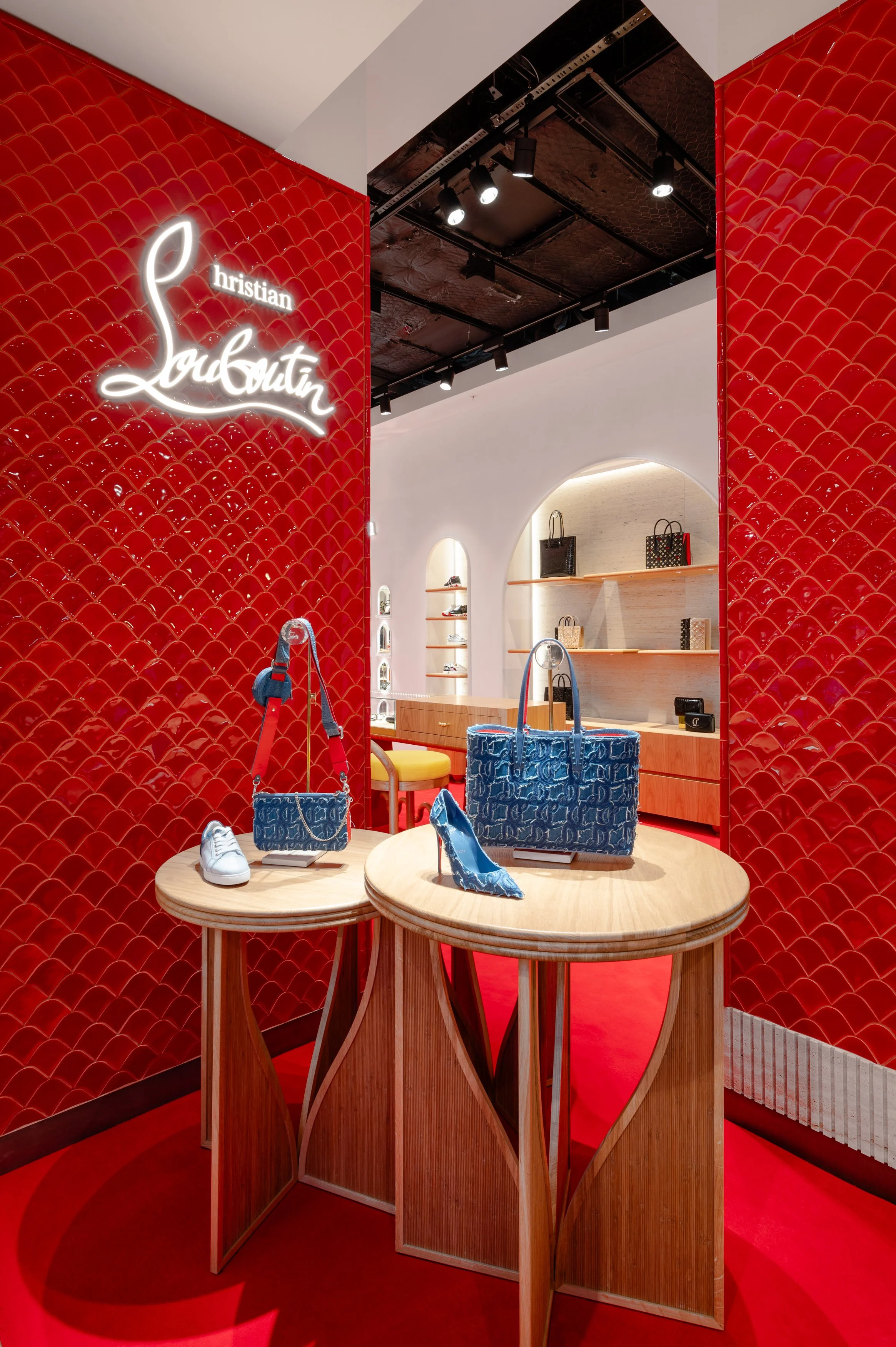 Christian Louboutin display inside a store with red patterned walls, wooden tables showcasing shoes, handbags, and accessories, and black ceiling lights.
