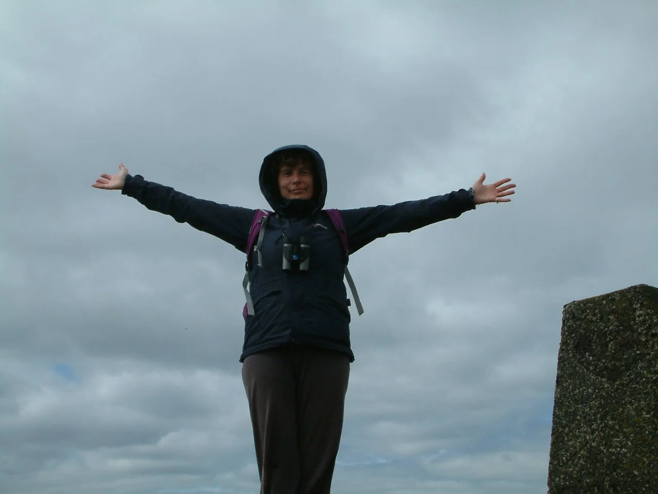 A person standing on a mountain with arms outstretched, wearing a jacket and backpack, under a cloudy sky.