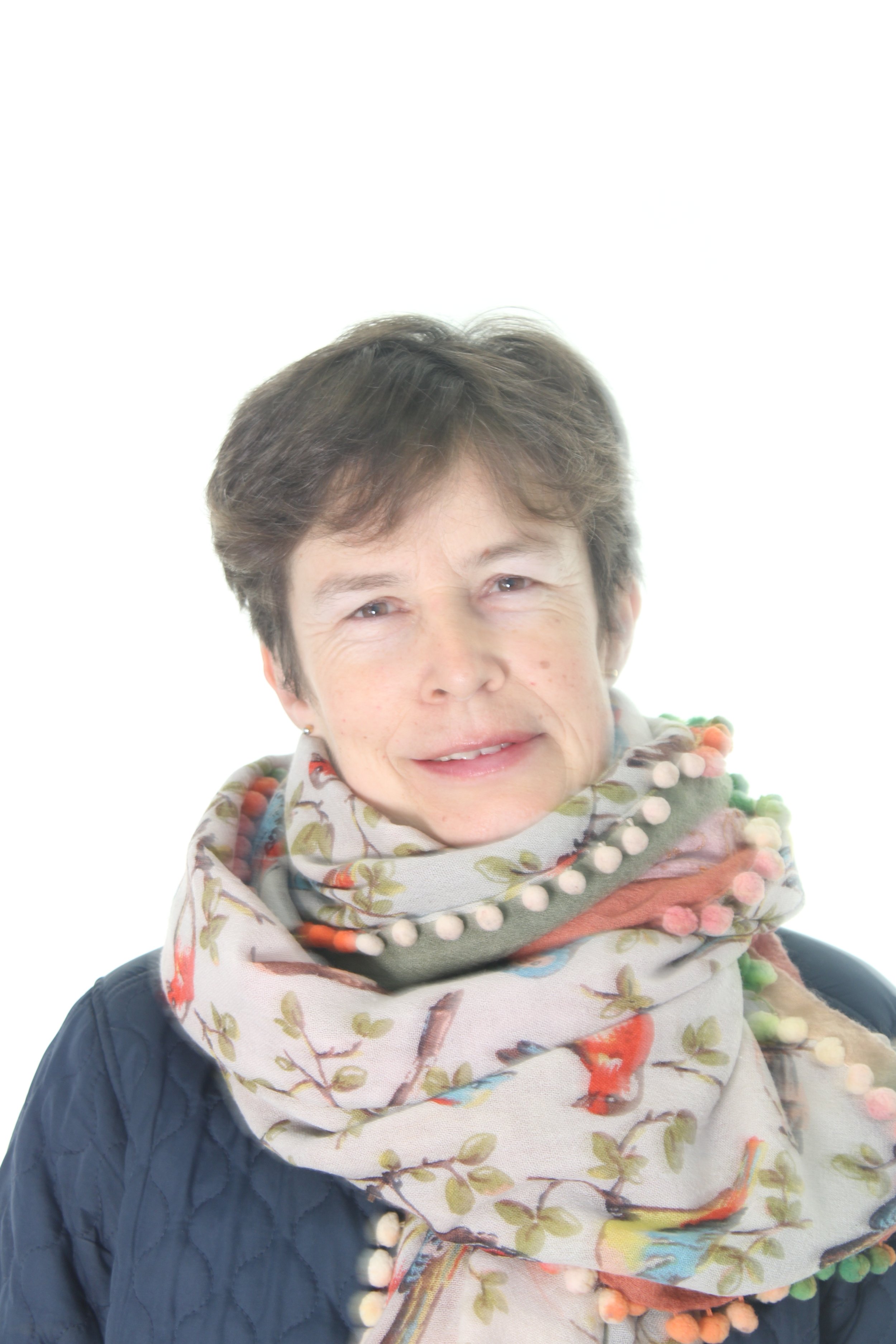 A portrait of a woman with short brown hair, smiling, wearing a colorful, patterned scarf with pom-poms and a dark quilted jacket, against a white background.