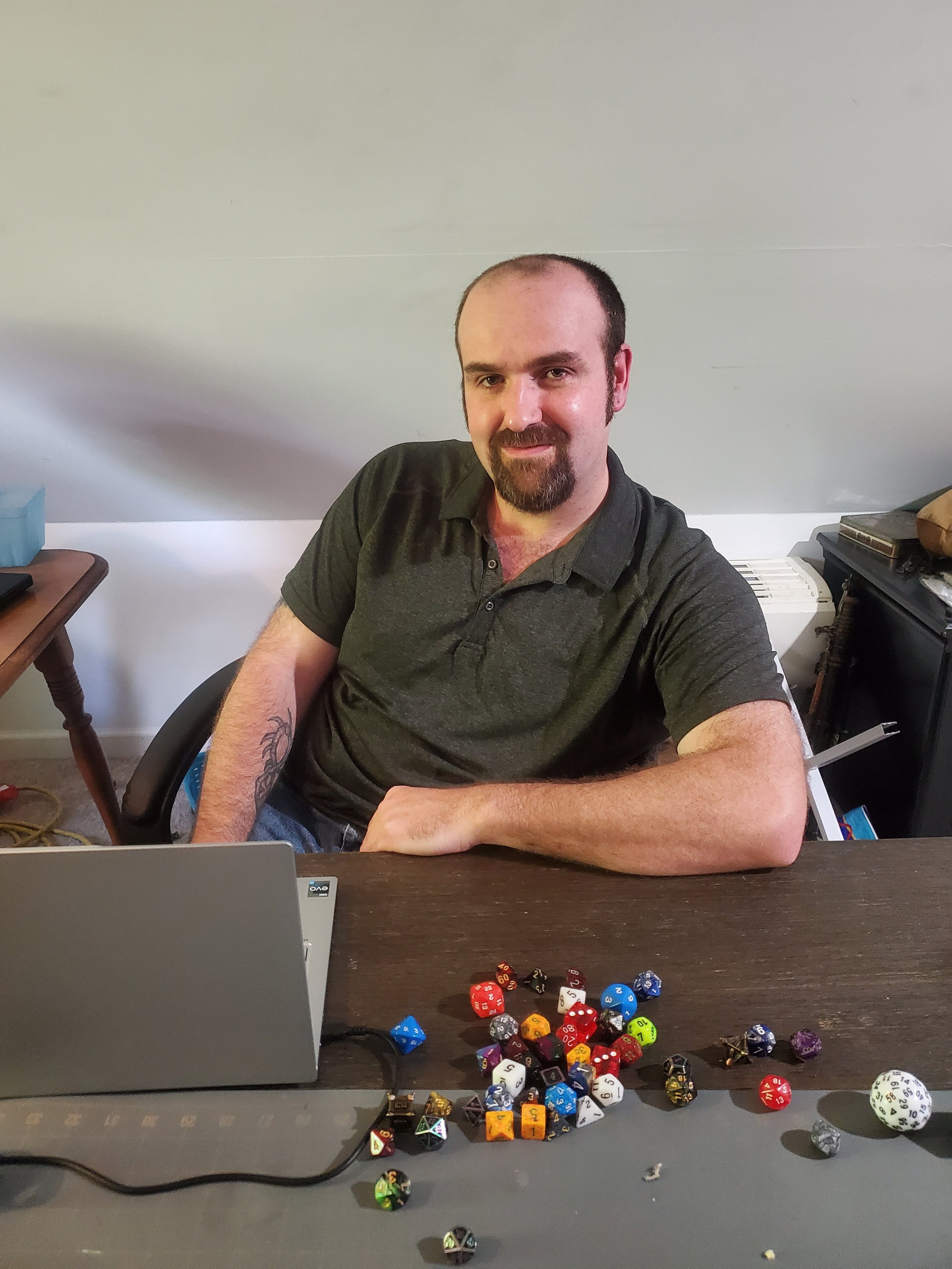 A man with a goatee and short hair, wearing a dark polo shirt, sitting at a desk with various colorful gaming dice and a laptop in front of him, smiling at the camera.
