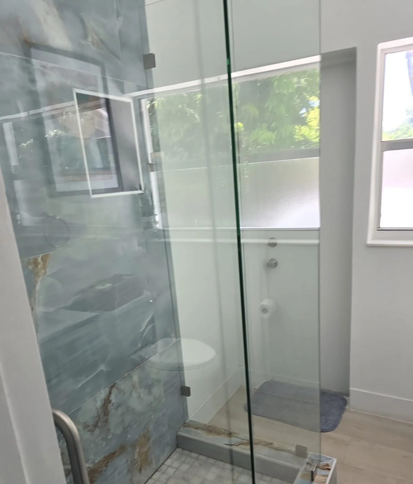 Glass shower enclosure with marble or tile walls, viewed through a glass door. The bathroom has a window with greenery visible outside, and a small gray bath mat on the floor.