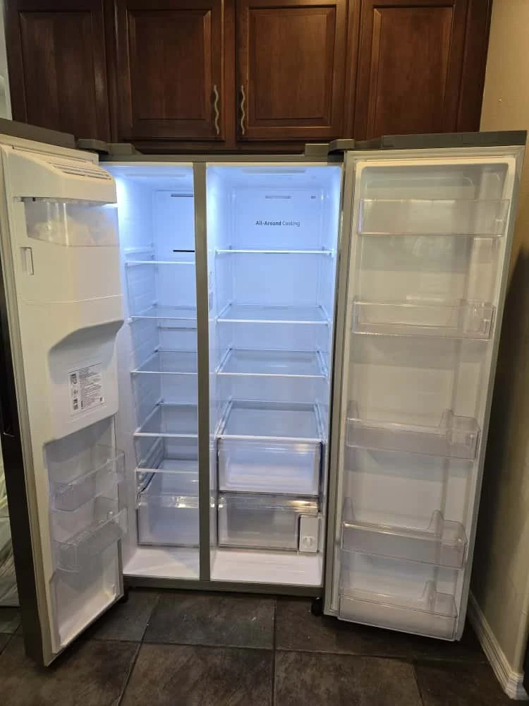 An empty side-by-side refrigerator with the doors open, showing the interior and shelves, situated next to kitchen wooden cabinets.