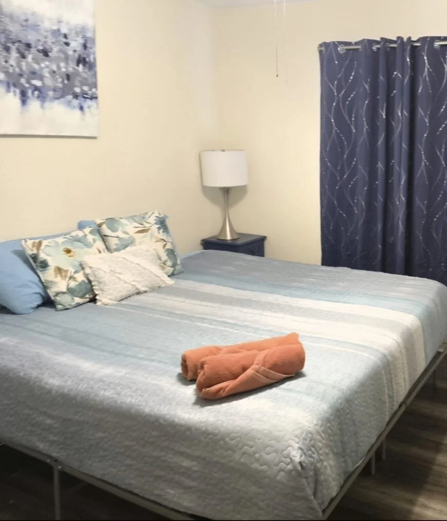 A neatly made bed with a blue and floral quilt, several pillows, and a folded blanket shaped like a dog. There is a nightstand with a lamp next to the bed, a blue curtain with a wavy pattern, and a partial abstract painting on the wall.
