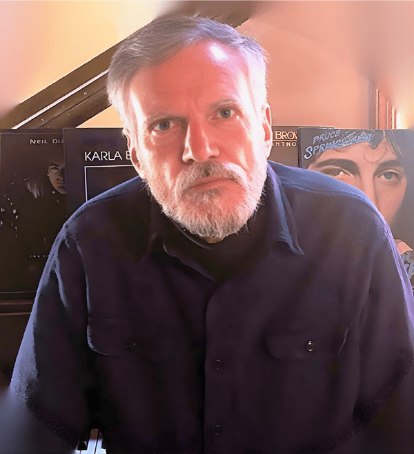 Author Peter Sarno with gray hair, beard, and serious expression sitting in front of a piano with posters of albums or book covers behind him.