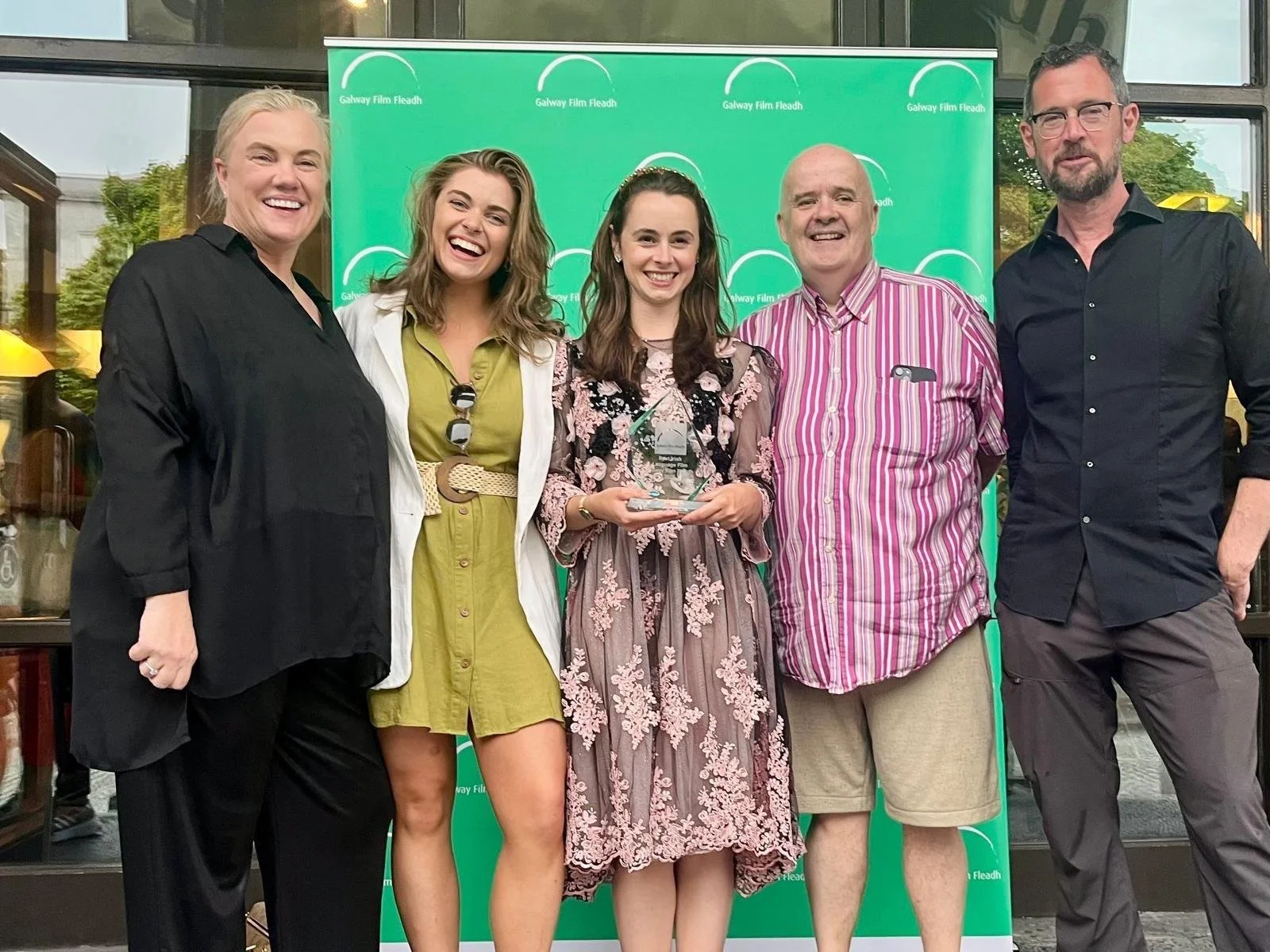 Group of six people standing in front of a green background with text, smiling, with one woman in the center holding an award or trophy.