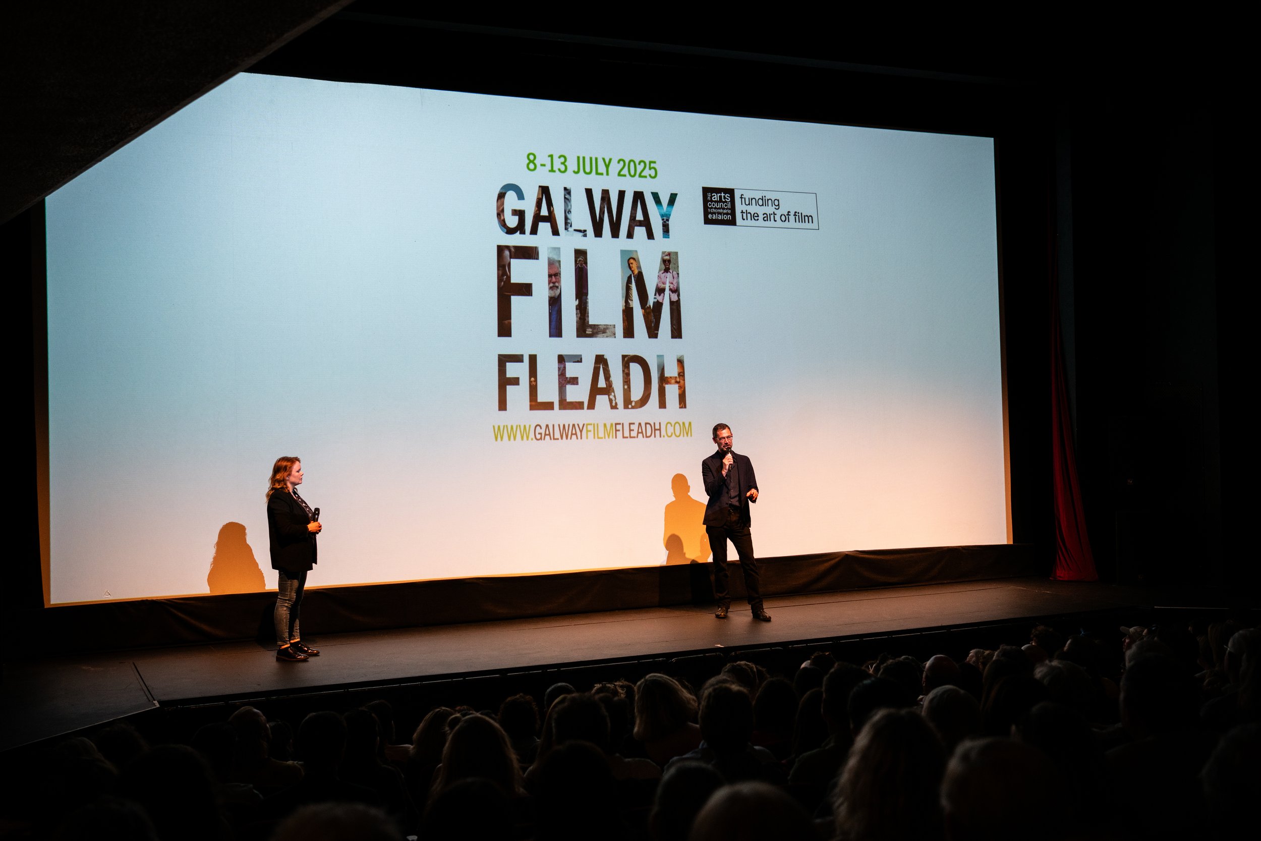 Two presenters on stage at the Galway Film Fleadh film festival, with a large screen displaying the event details and website URL in the background.