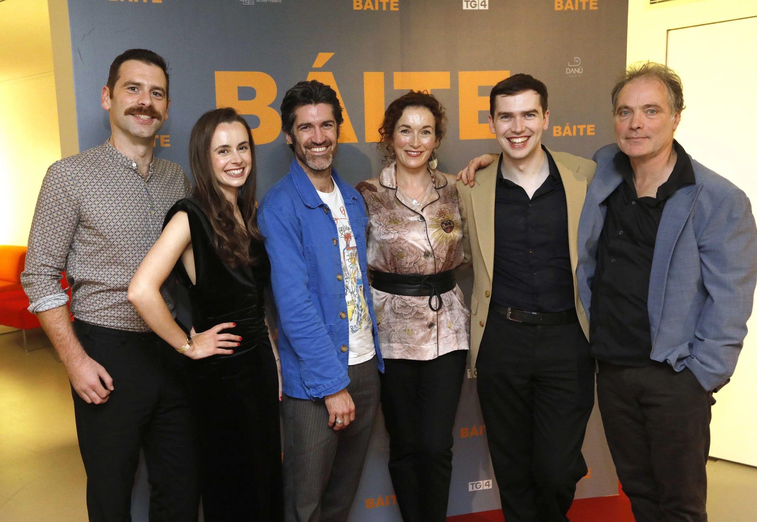 Group of six smiling people standing together in front of a dark gray backdrop with the word 'BÁITE' repeatedly printed in large orange letters. They are indoors, dressed in casual and semi-formal attire, with a yellow wall on the right and an orange chair partially visible on the left.