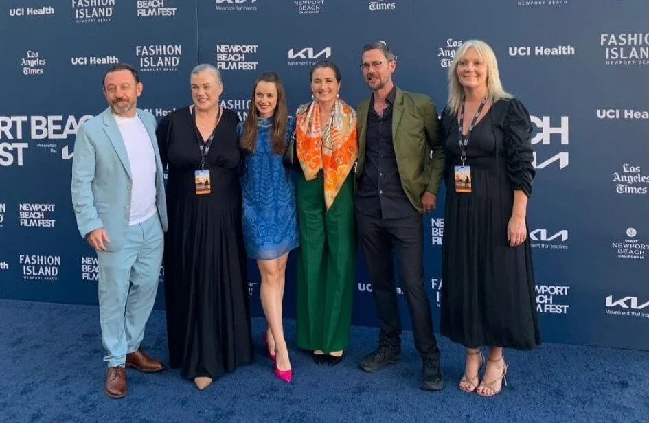 Six people standing side by side on a blue carpet at an event with a blue backdrop featuring logos and names such as 'Fashion Island,' 'Newport Beach Film Fest,' and 'Los Angeles Times.' They are smiling and dressed in formal and semi-formal attire.