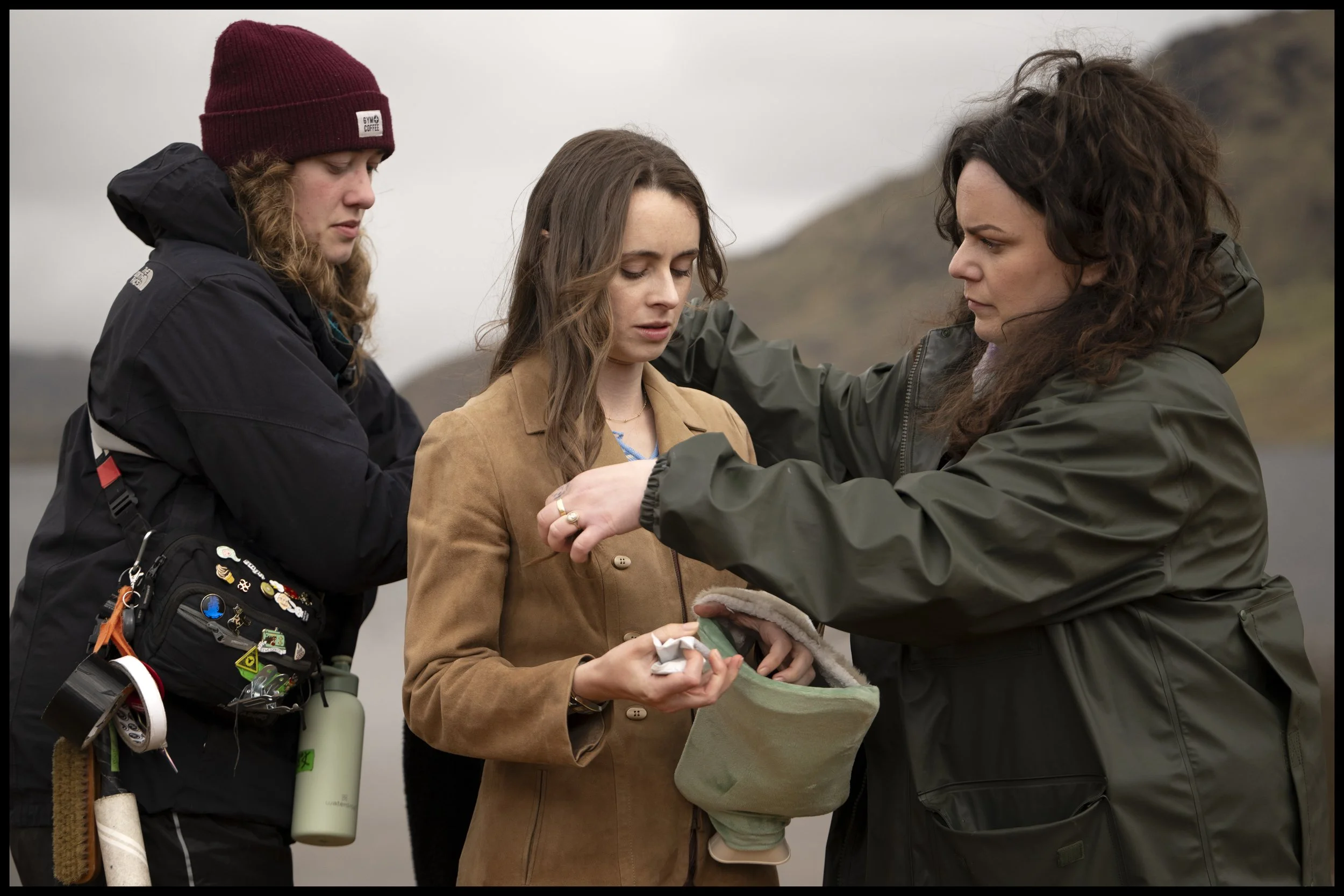 Three women outdoors in a rural, mountainous area during cloudy weather. One woman is adjusting another woman's jacket while a third looks on, all dressed in outdoor gear.