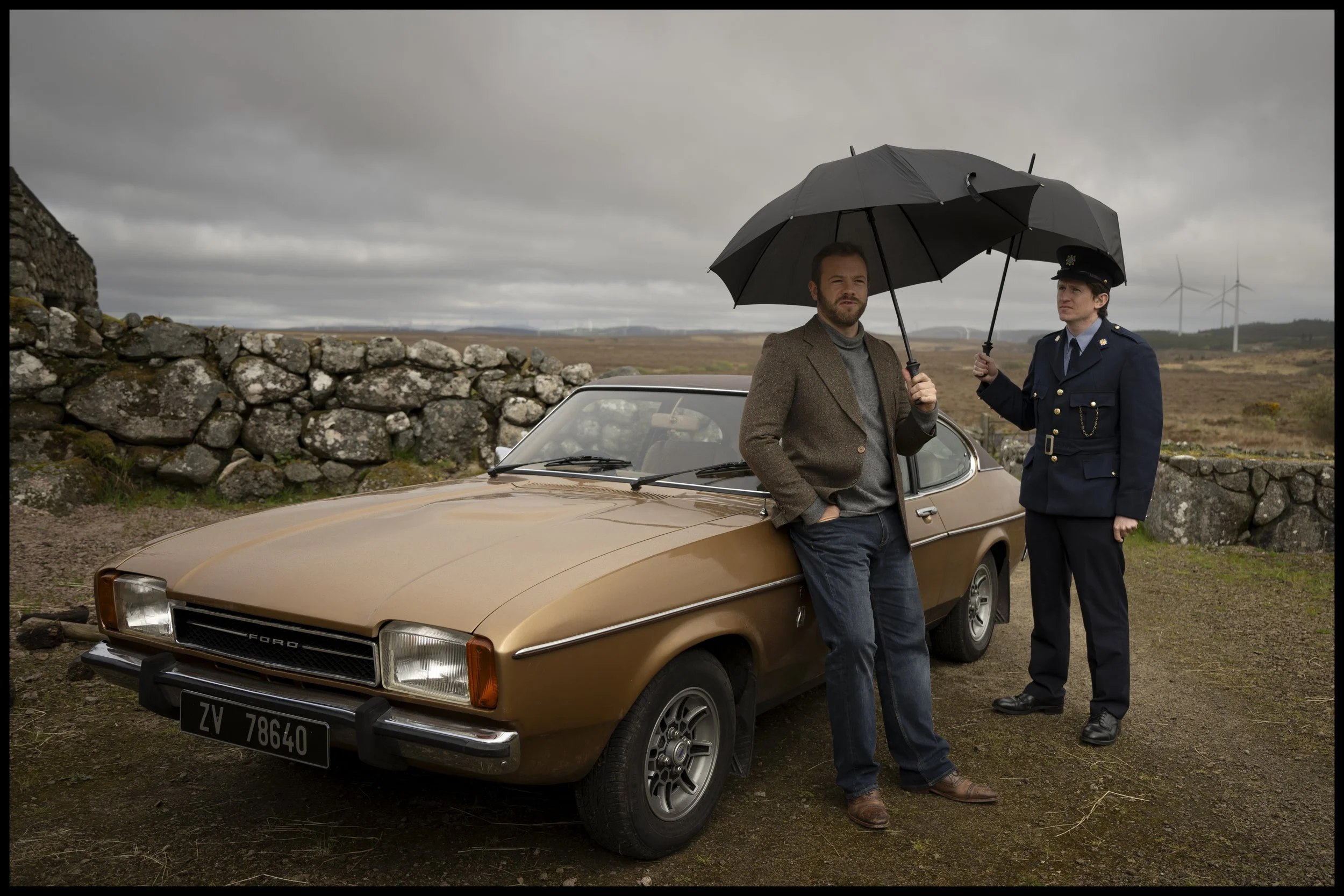 A man leaning against a vintage brown Ford car and a woman in a police uniform holding an umbrella over both of them, standing outdoors near a stone wall on a cloudy day, with wind turbines in the background.