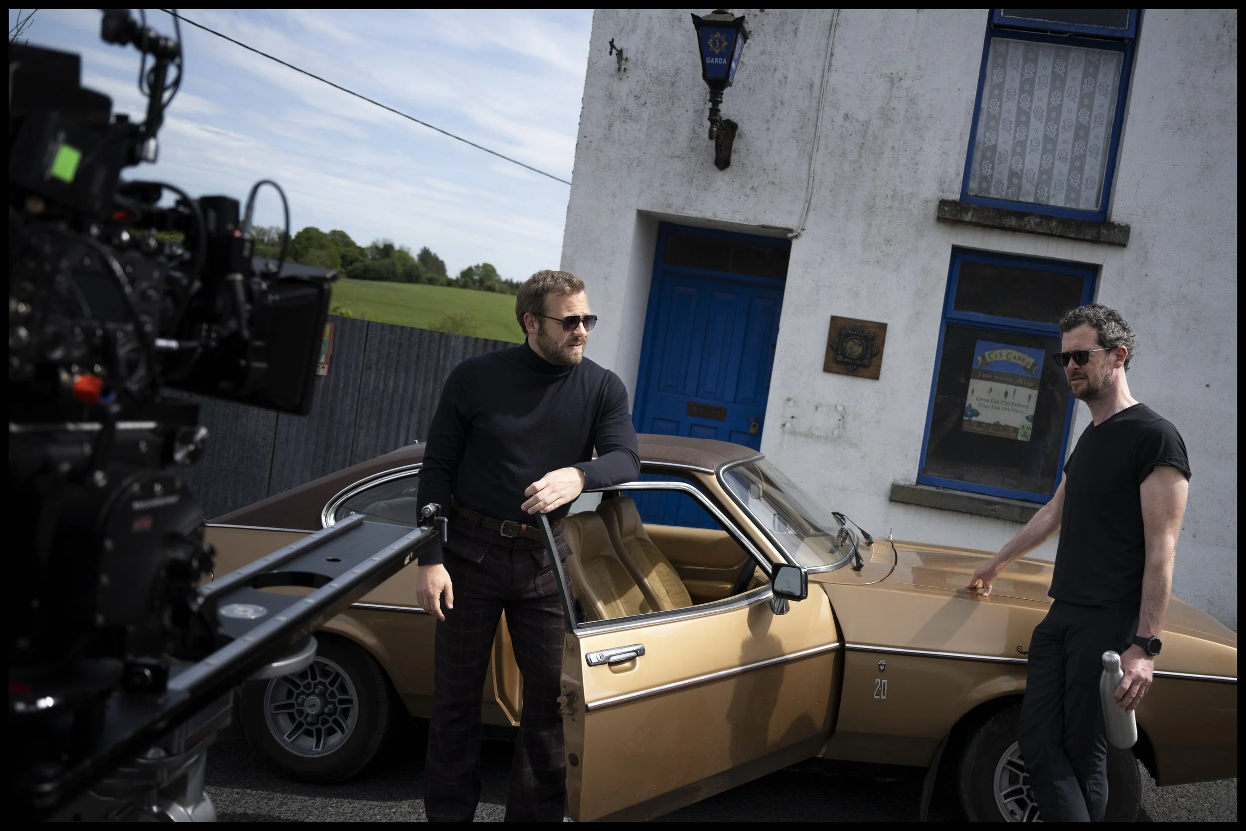 Two men with sunglasses stand next to a vintage gold-colored sports car with one arm resting on the open driver's door, outside a white building with blue trim and a blue door, while a camera records the scene.