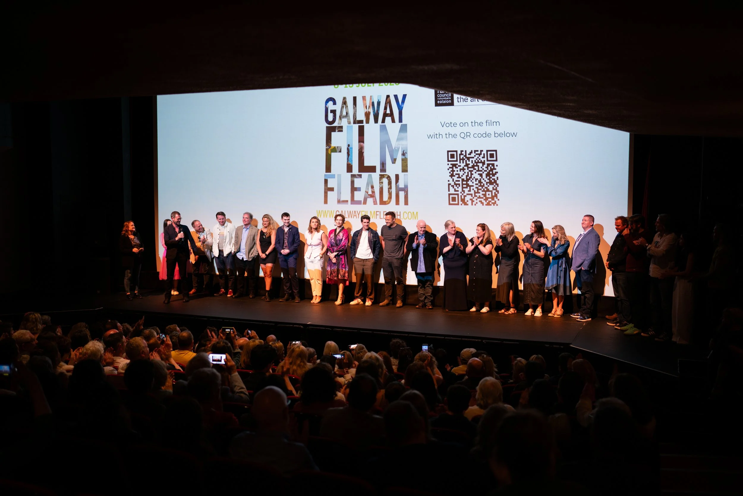 People standing on stage during an awards ceremony at Galway Film Fleadh, with a large screen in the background displaying event branding and a QR code, audience seated watching.
