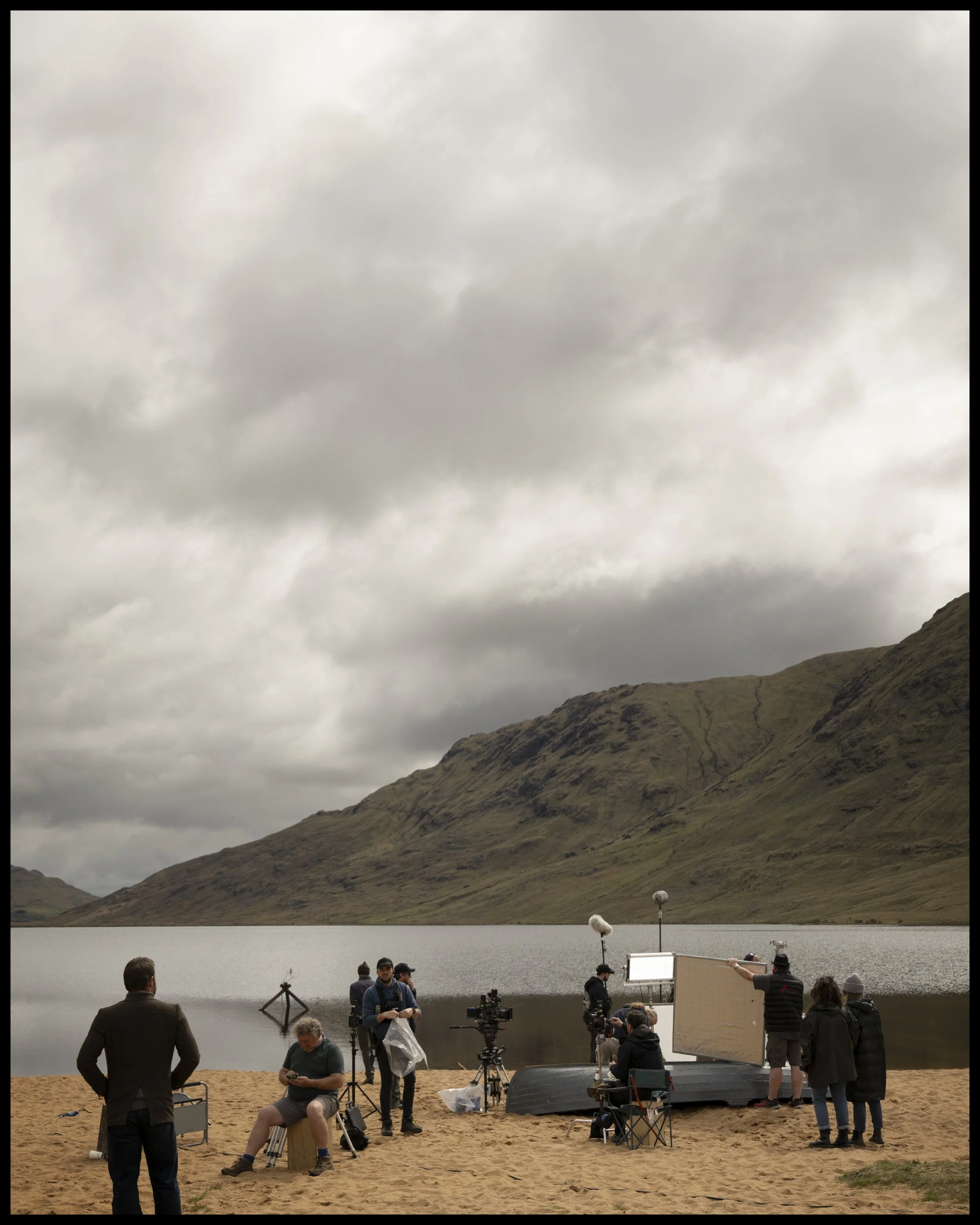 Film crew setting up equipment on a sandy beach with a lake and mountains in the background under overcast skies.