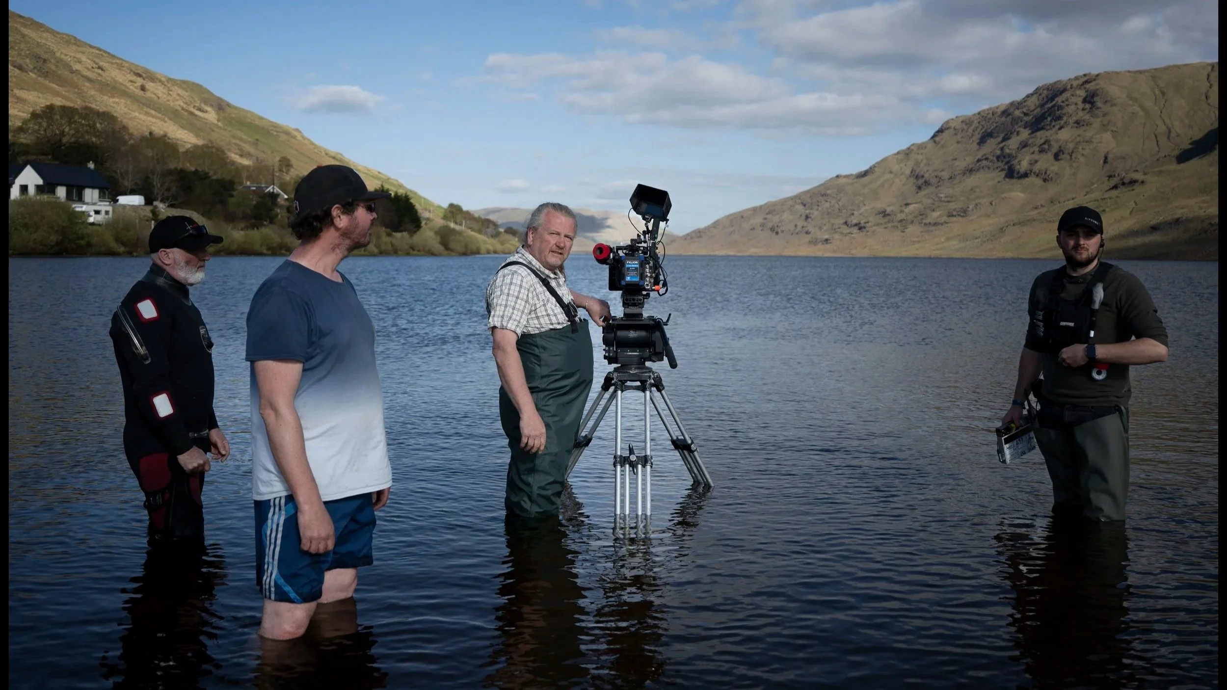 Group of five men filming a scene in a lake with mountains and a partly cloudy sky in the background.