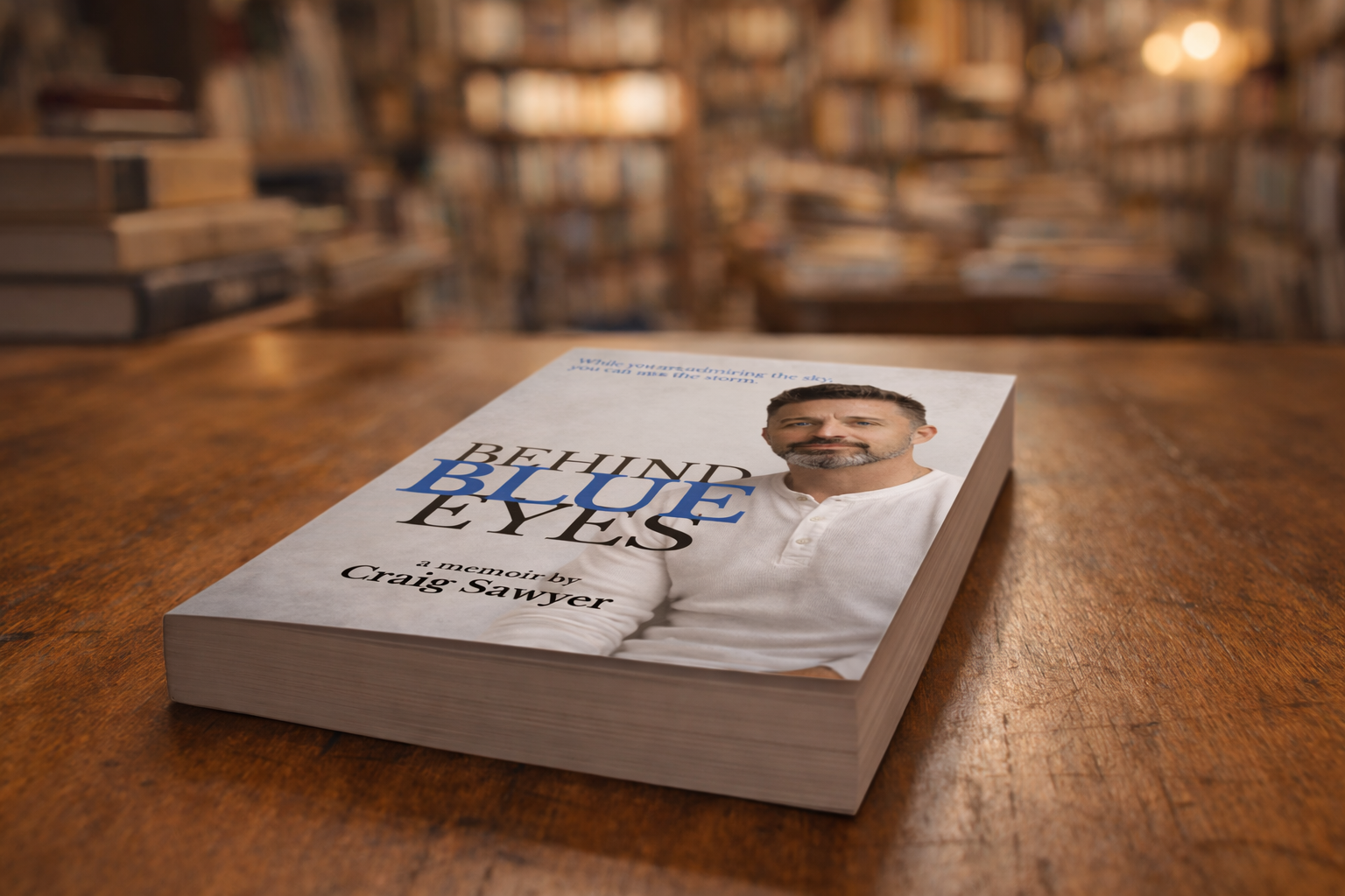 A paperback book titled 'Behind Blue Eyes' by Craig Sawyer lying on a wooden table in a bookstore with shelves full of books in the background.