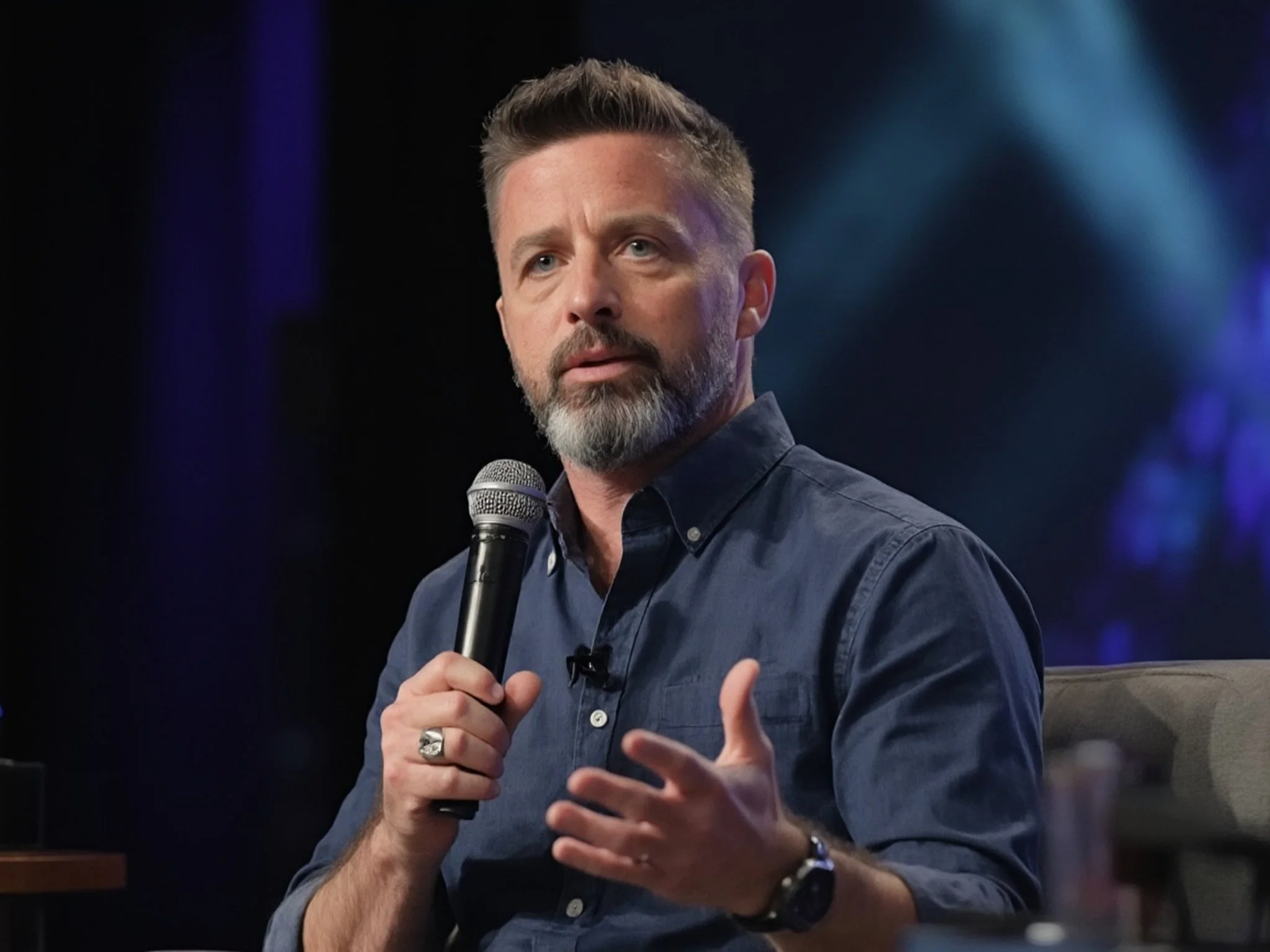 Man with a beard and short hair, wearing a dark blue shirt, holding a microphone in his right hand, sitting on a stage during a talk or panel discussion.