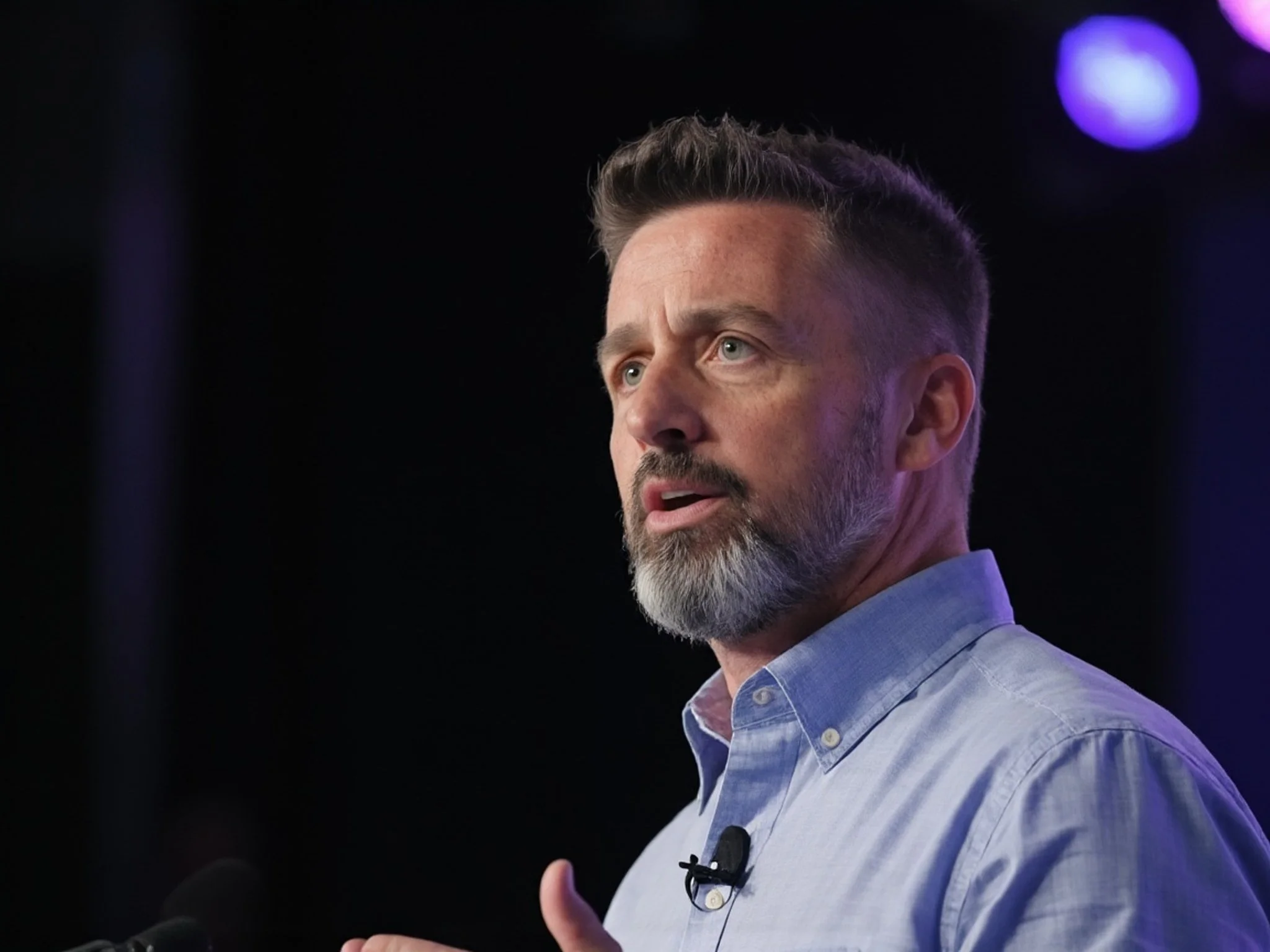 A middle-aged man with a beard and short hair, wearing a light blue button-up shirt, is speaking or giving a presentation against a dark background with purple lights.