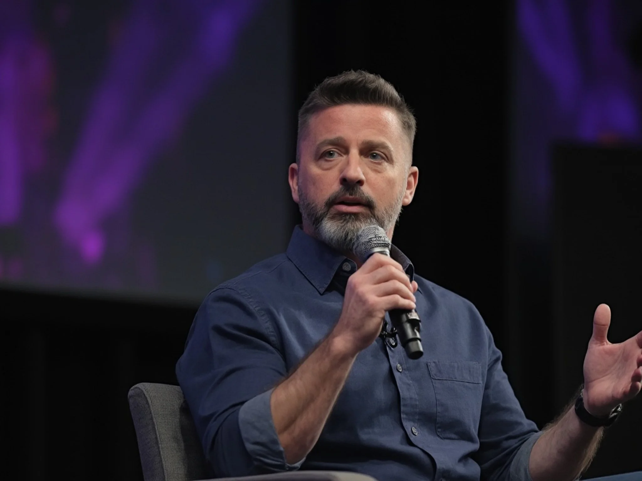 A man with a beard and short hair is sitting on a chair, speaking into a microphone during an event or conference, with dark background and purple lighting.