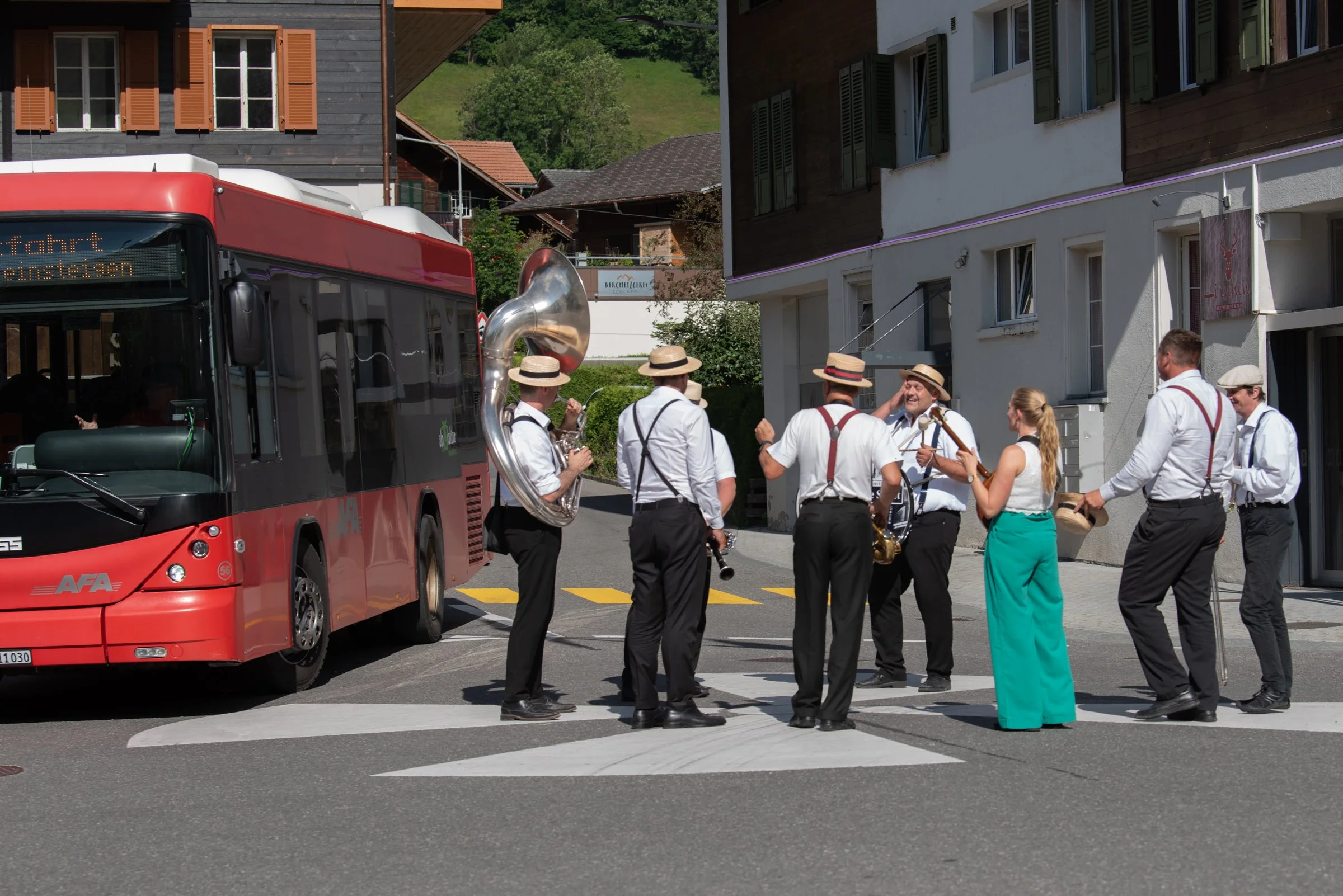 Eine Gruppe von Musikern in weißen Hemden und Hüten, die auf einer Straßenkreuzung ein Straßenkonzert mit Instrumenten wie Trompete, Saxophon, Klarinette und Tuba geben, während ein roter Bus vorbeifährt.