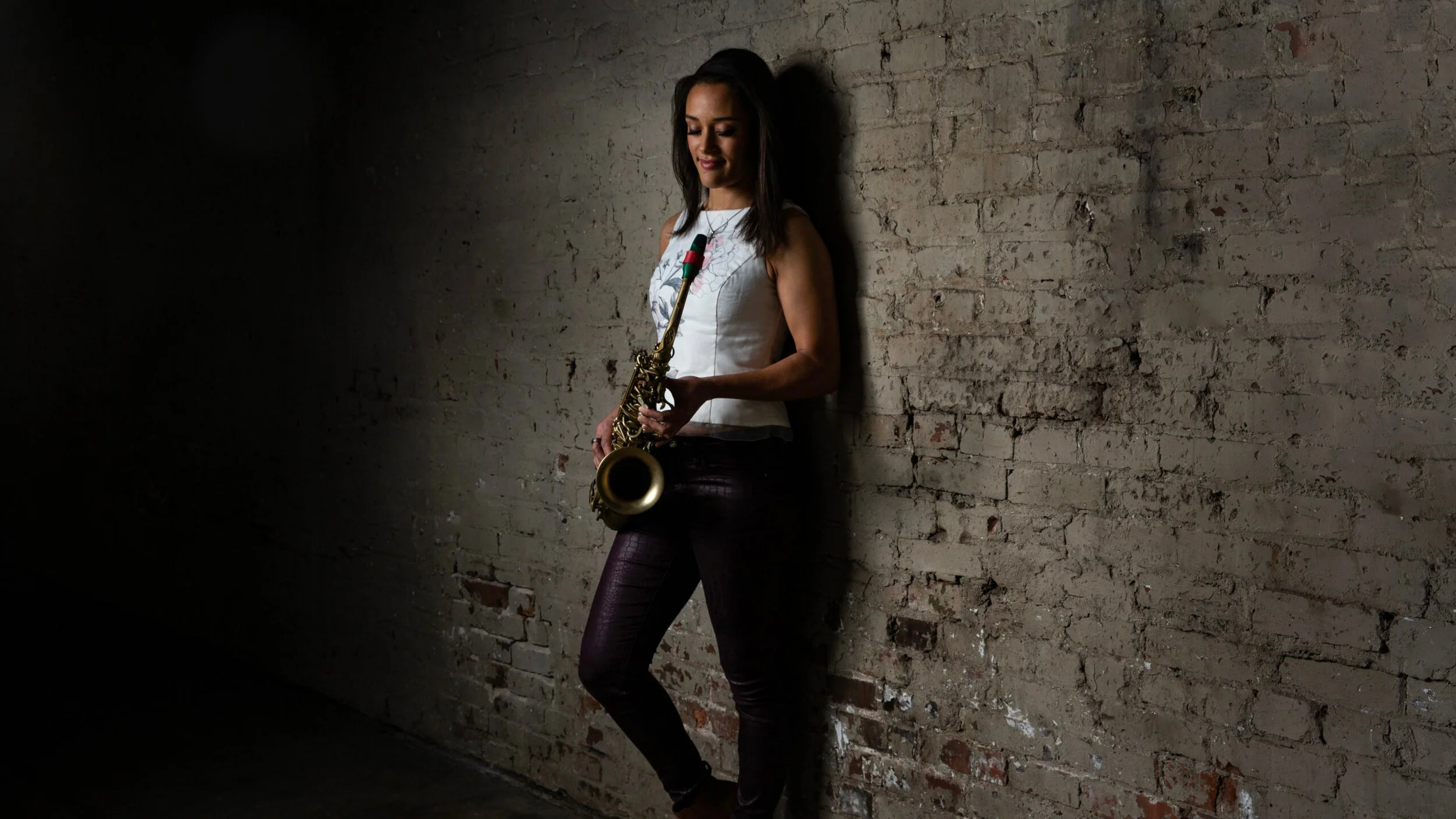 Junge Frau mit einem Saxophon lehnt an einer alten Backsteinmauer in einem dunklen Raum.
