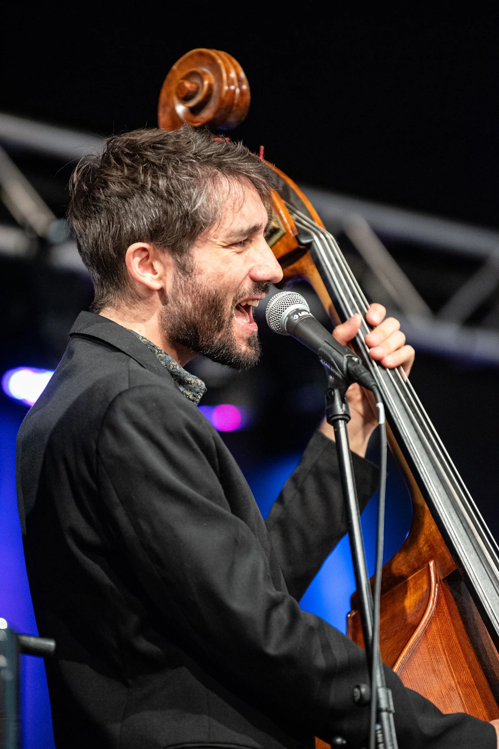 Ein männlicher Musiker singt, während er ein Kontrabass spielt und vor einem Mikrofon steht, auf einer Bühne bei einem Live-Auftritt.