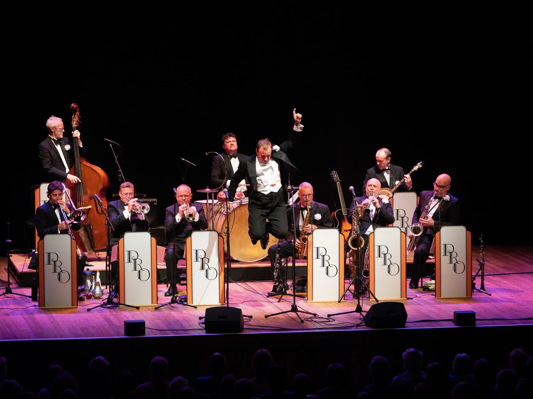 Jazzband mit zehn Musikern auf Bühne, einer springt in die Luft, alle in Anzügen, mit klassischen Jazzinstrumenten wie Trompete, Saxophon, Trommel, Bass, Gitarre und Mikrofon, vor dunklem Hintergrund bei Konzert.