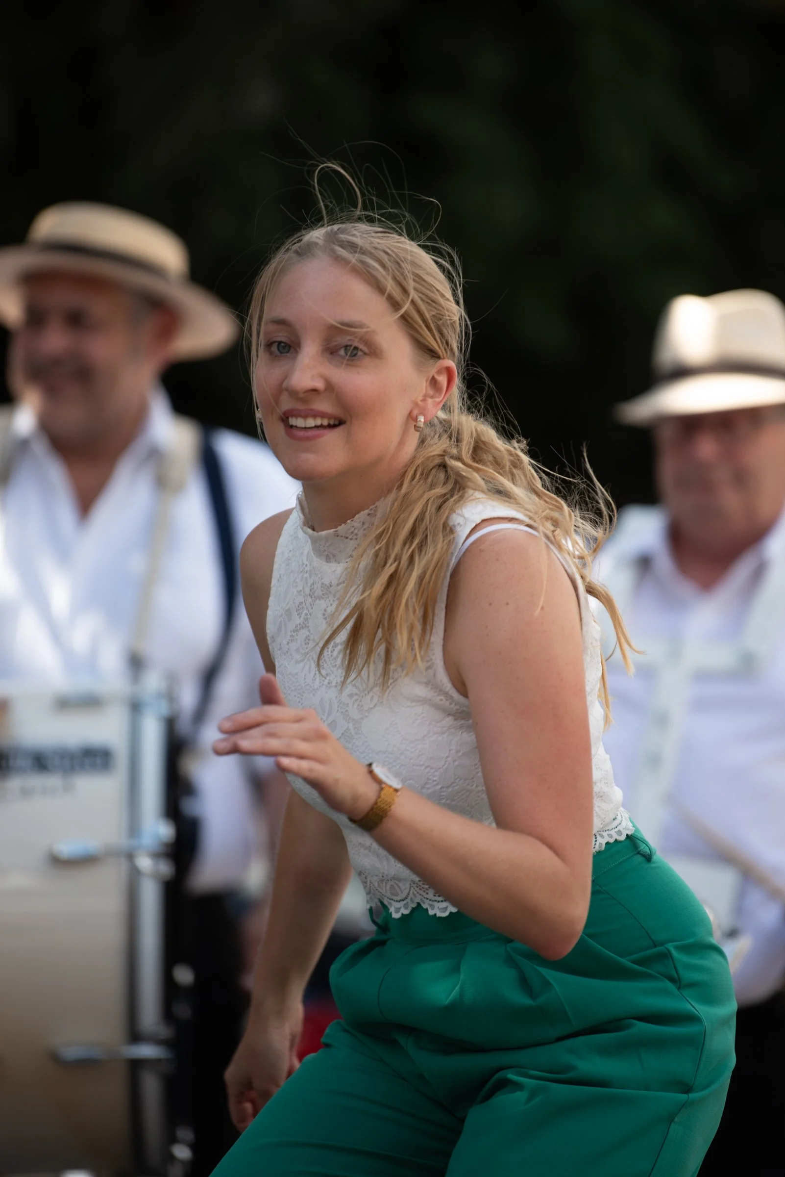 Junge Frau in weißer Spitzenbluse und grünen Hosen, tänzerisch auf einer Party im Freien, im Hintergrund Musiker mit Hüten.