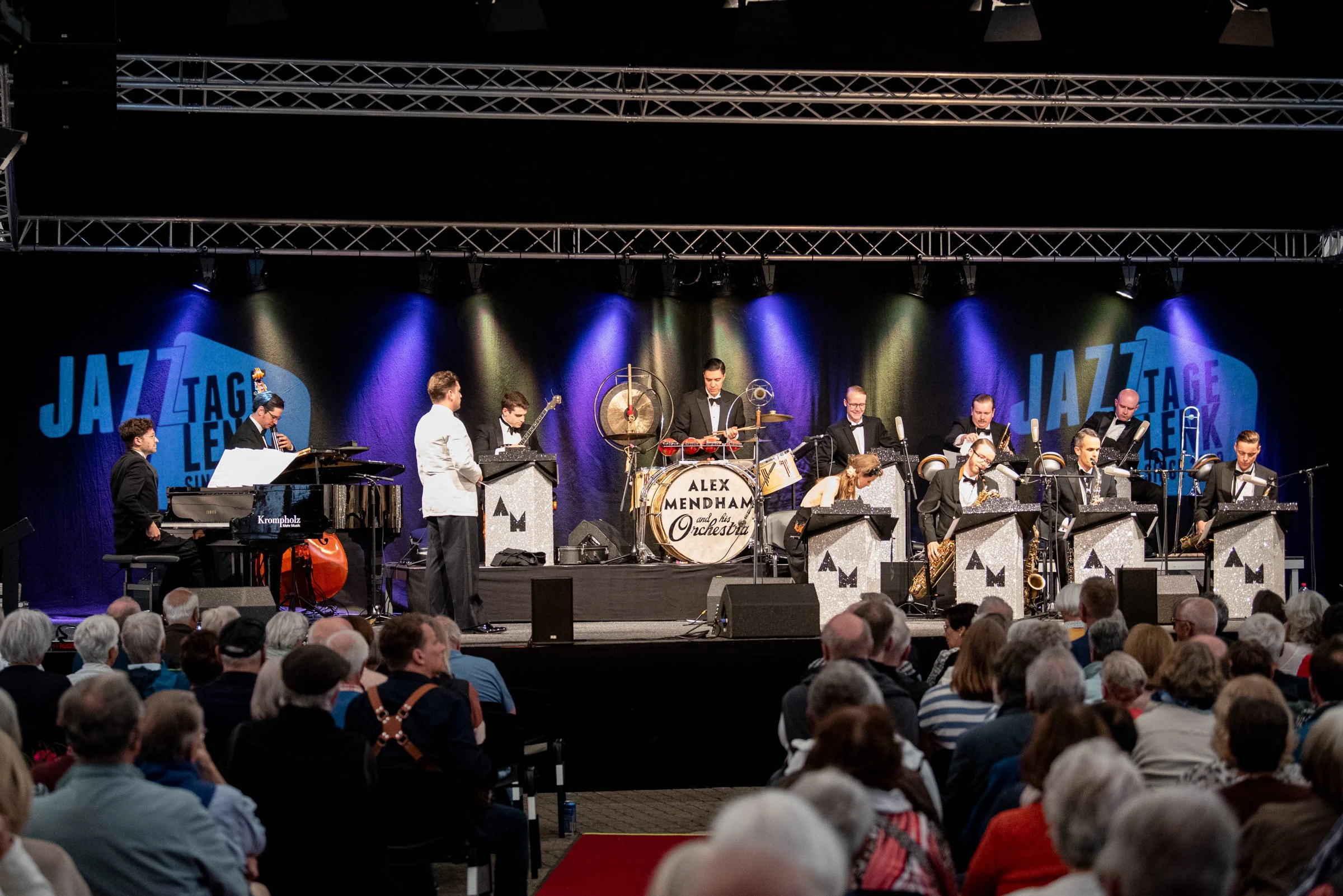 Ein Jazzorchester auf einer Bühne bei einem Konzert, mit mehreren Musikern, die an Instrumenten wie Klavier, Bass, Schlagzeug, Saxophon und Trompete spielen. Das Bühnenhintergrund ist schwarz mit blauen Beleuchtungen und einem Logo für das Jazzfestiv