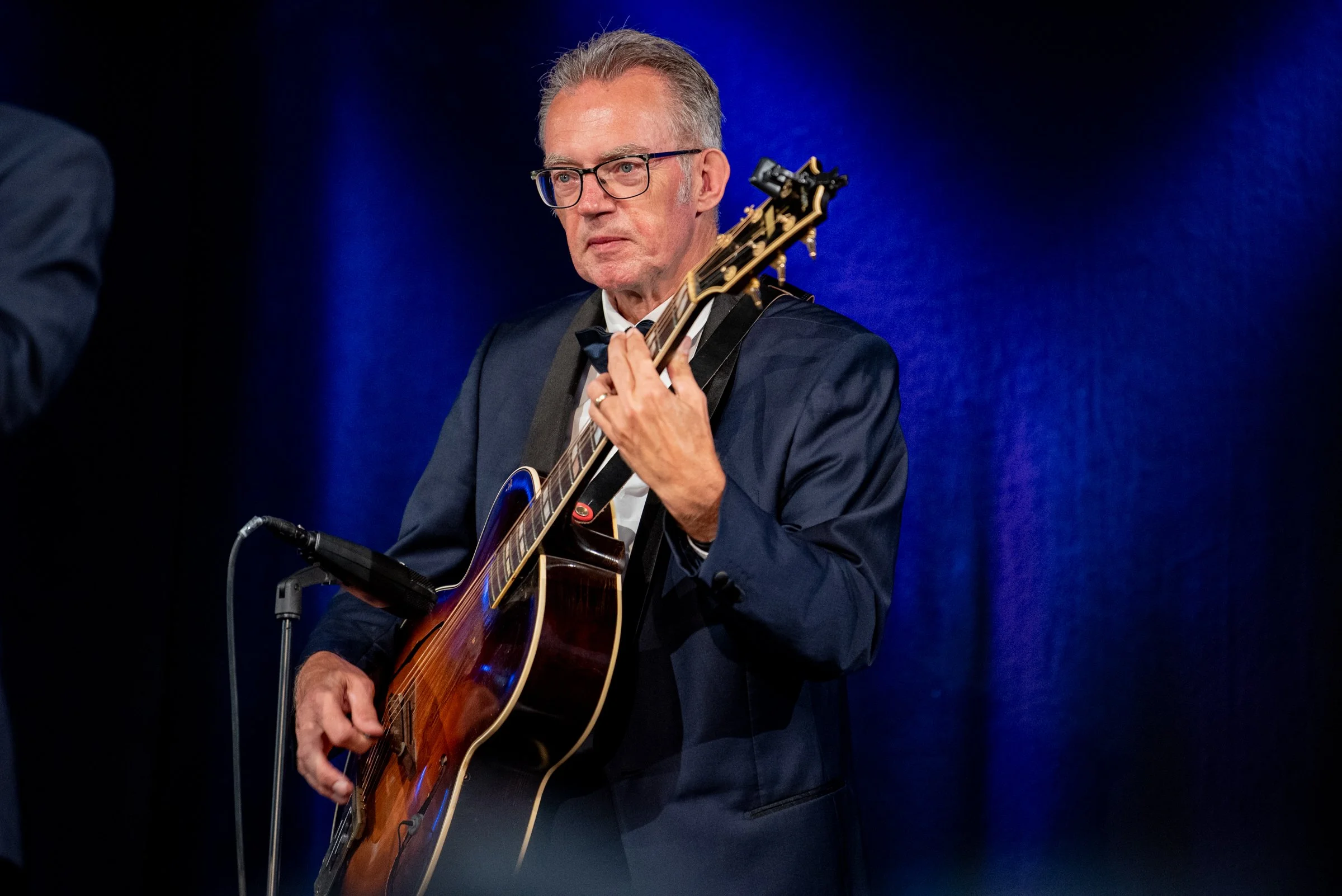 Ein älterer Mann in formeller Kleidung spielt Gitarre auf einer Bühne mit blauer Kulisse.