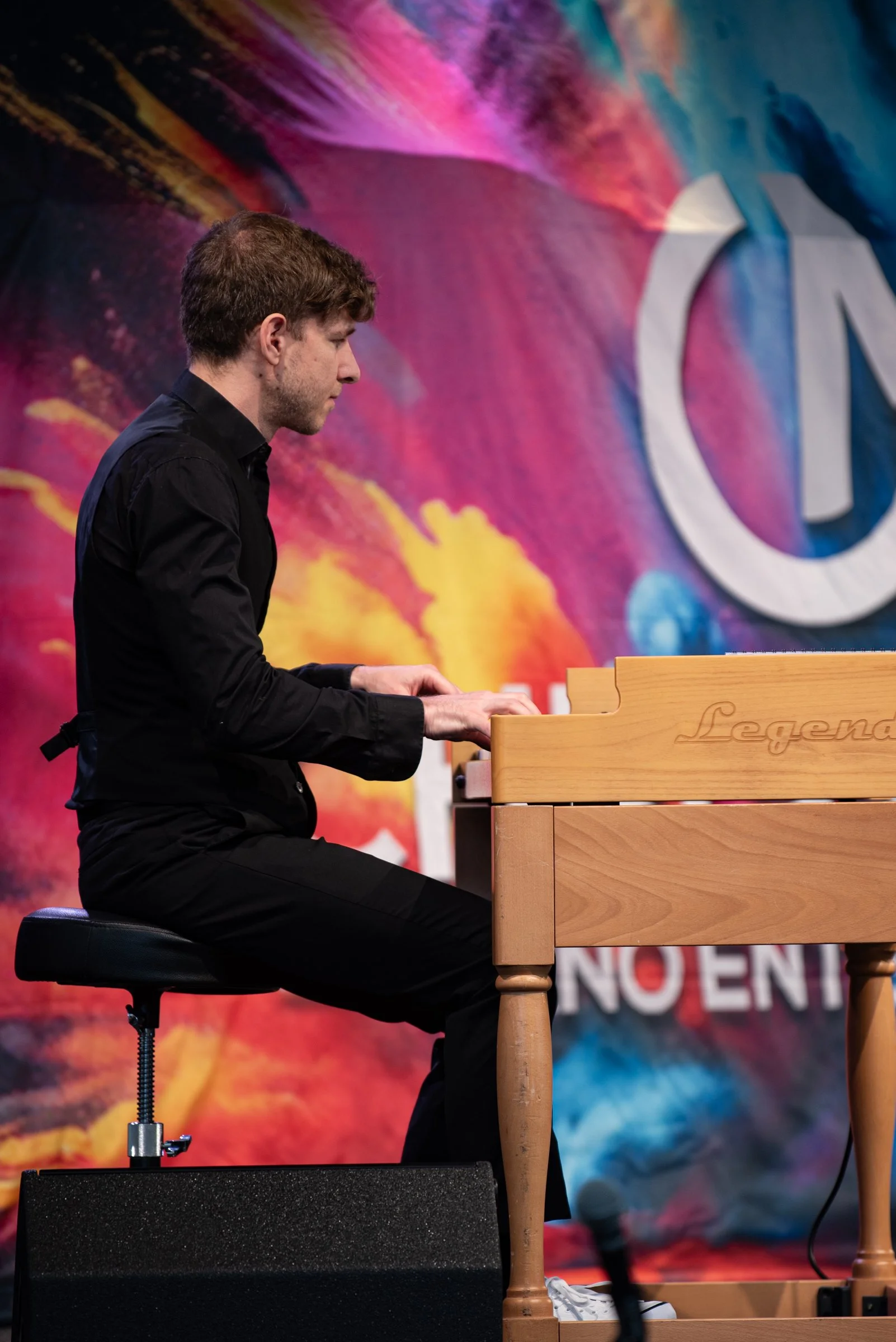 Junger Mann spielt Klavier auf einer Bühne mit bunter, abstrakter Hintergrundwand.