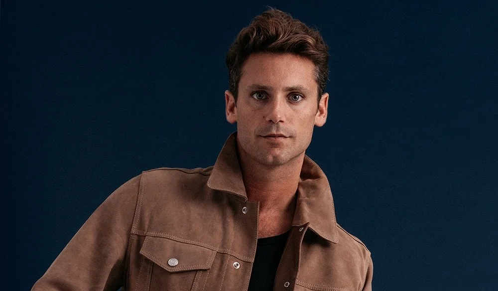 Ein junger Mann mit braunem Haar, trägt eine braune Jacke vor einem dunklen Hintergrund.