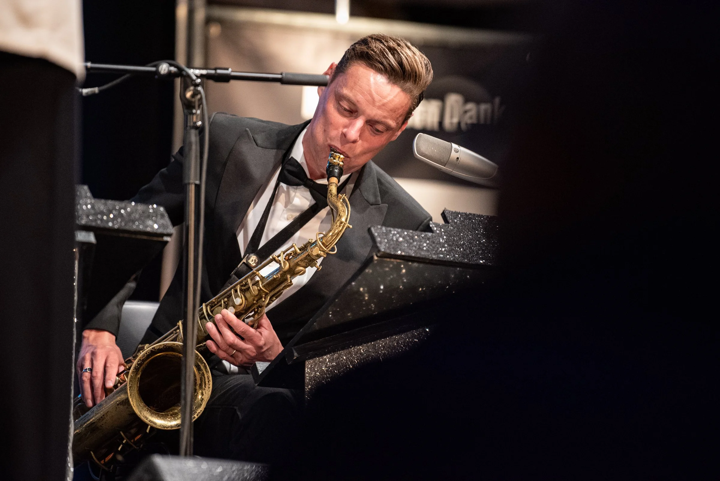 Ein Mann trägt einen schwarzen Smoking mit Fliege und spielt Saxophon beim Auftritt in einem dunklen Raum.