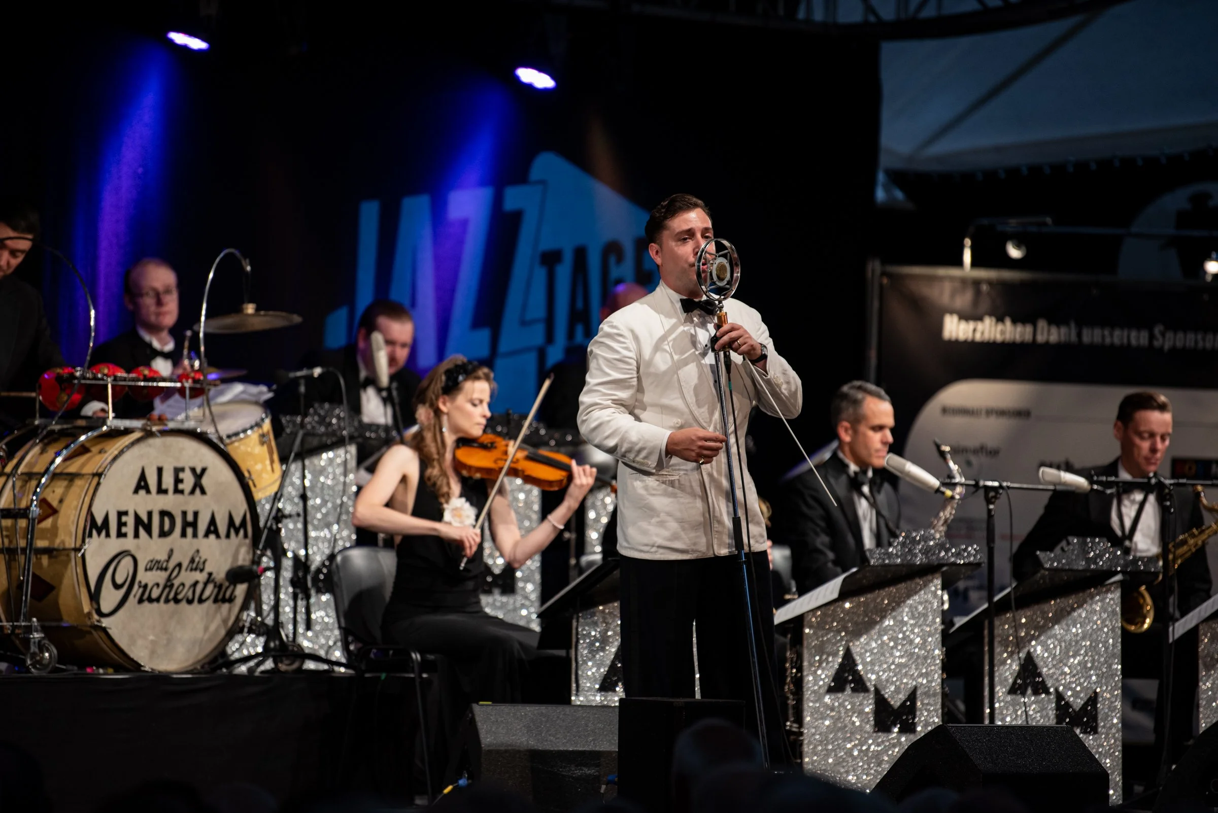 Musiker auf einer Bühne bei einem Open-Air-Konzert, eine Person singt oder spricht in ein Mikrofon, im Hintergrund sind weitere Musiker mit Instrumenten, einschließlich Schlagzeug, Geige, Saxophon und weiteren, mit Glitzer- und Schwarz-Weiß-Designs b