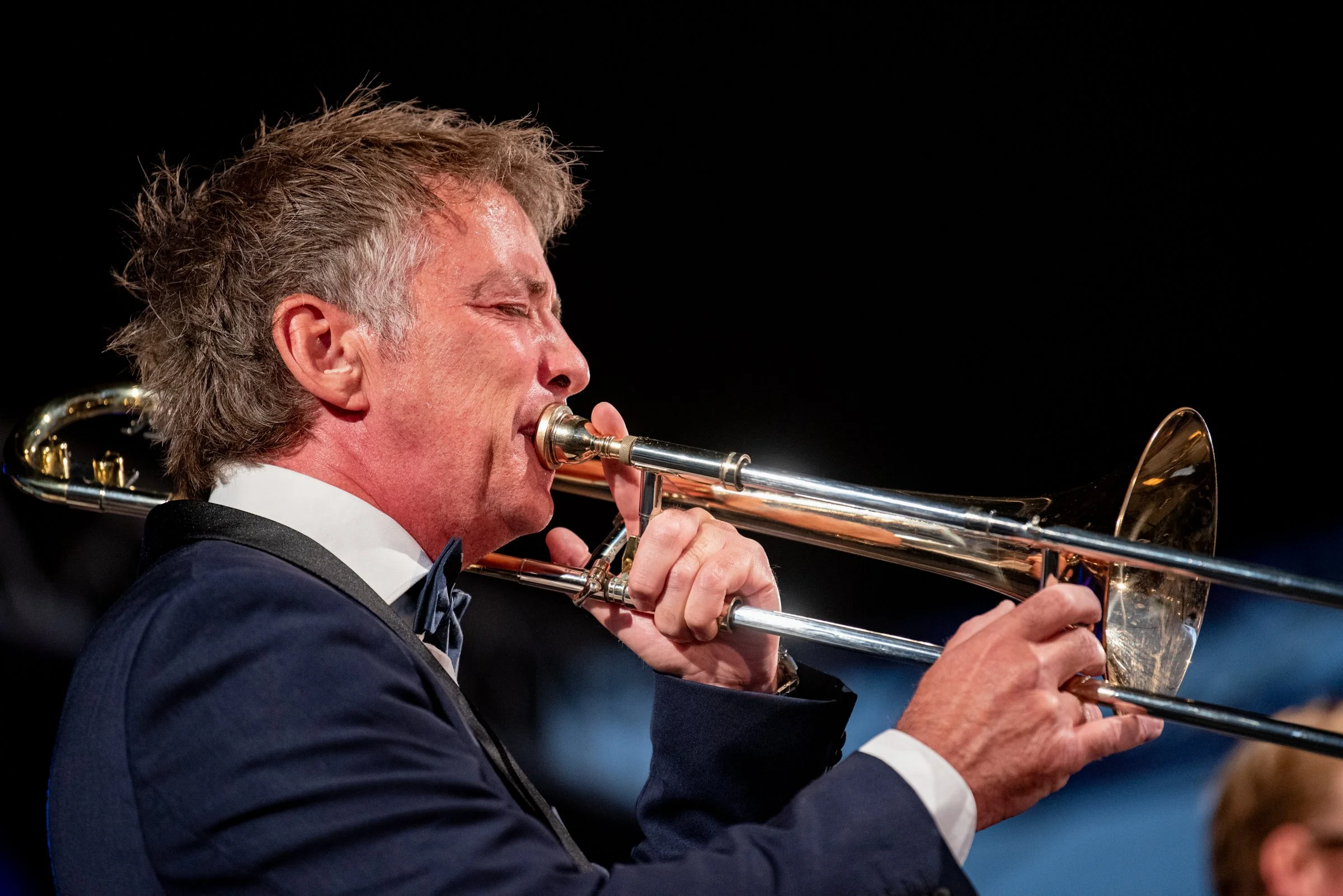 Ein Mann in einem schwarzen Anzug und Fliege spielt auf einer Trompete bei einem Konzert.