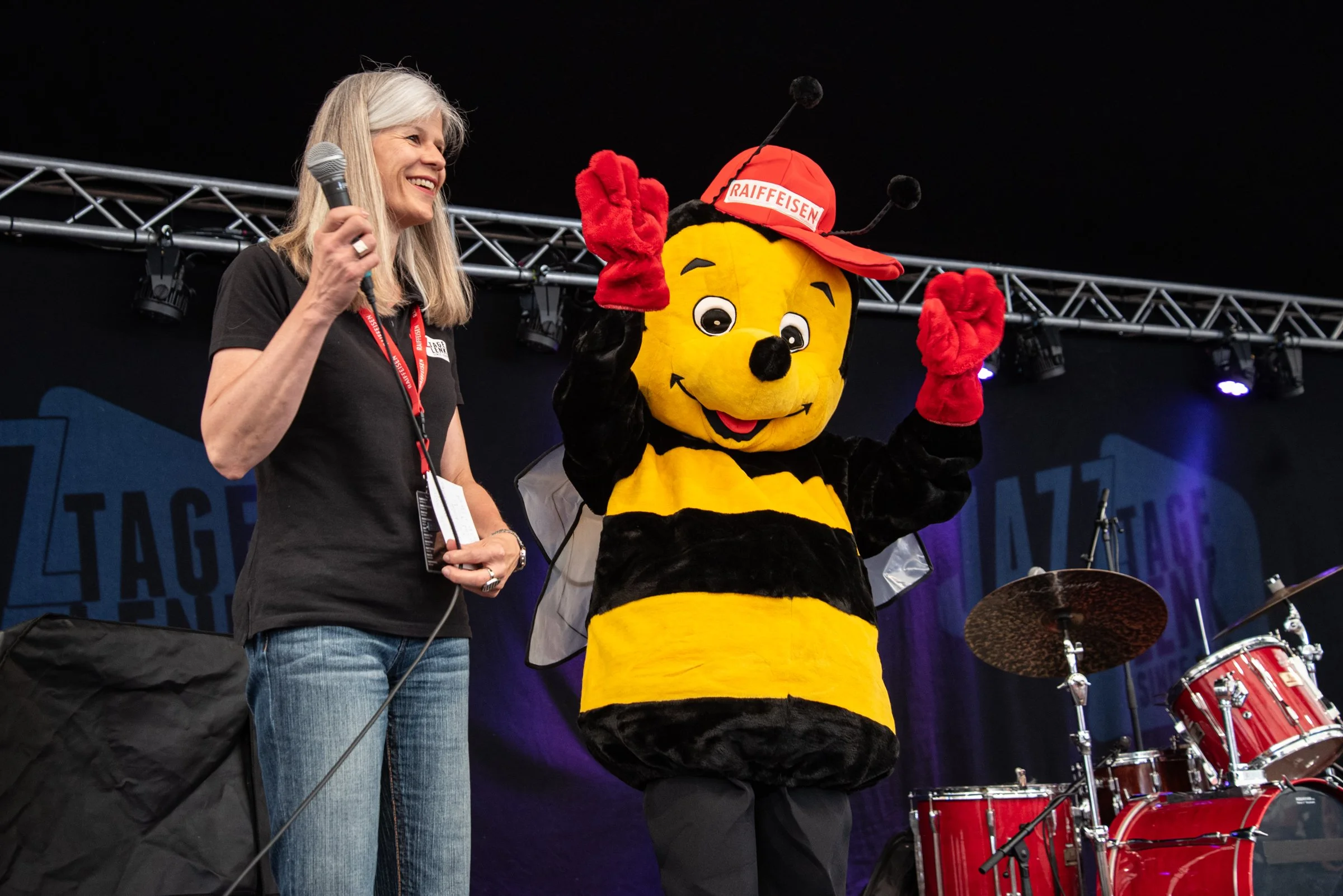Eine Frau hält ein Mikrofon und steht neben einer freundlichen Biene-Charakter in einem schwarz-gelben Anzug mit rotem Helm. Sie sind auf einer Bühne, im Hintergrund sind Lautsprecher und ein Schlagzeug zu sehen.