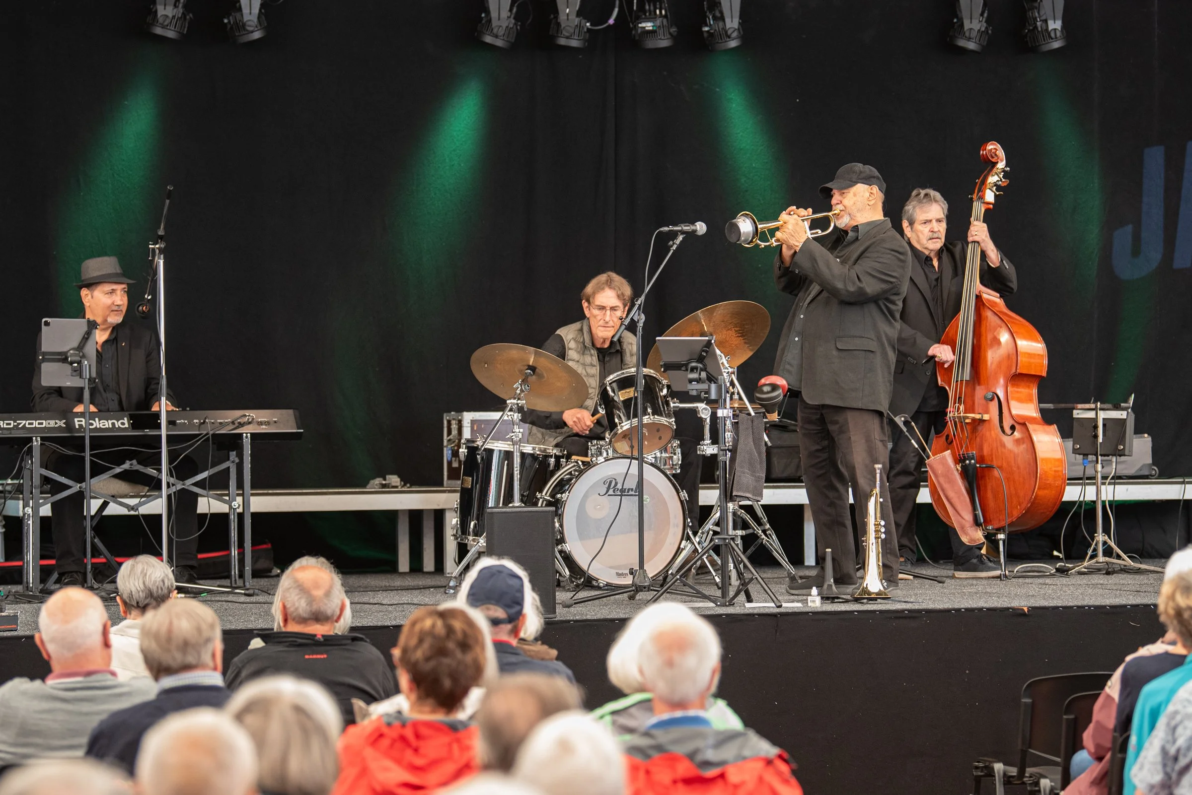 Jazzband auf einer Bühne beim Auftritt, mit einem Keyboarder, einem Drummer, einem Trompeter und einem Bassisten, im Publikum stehen Menschen, die das Konzert anschauen.