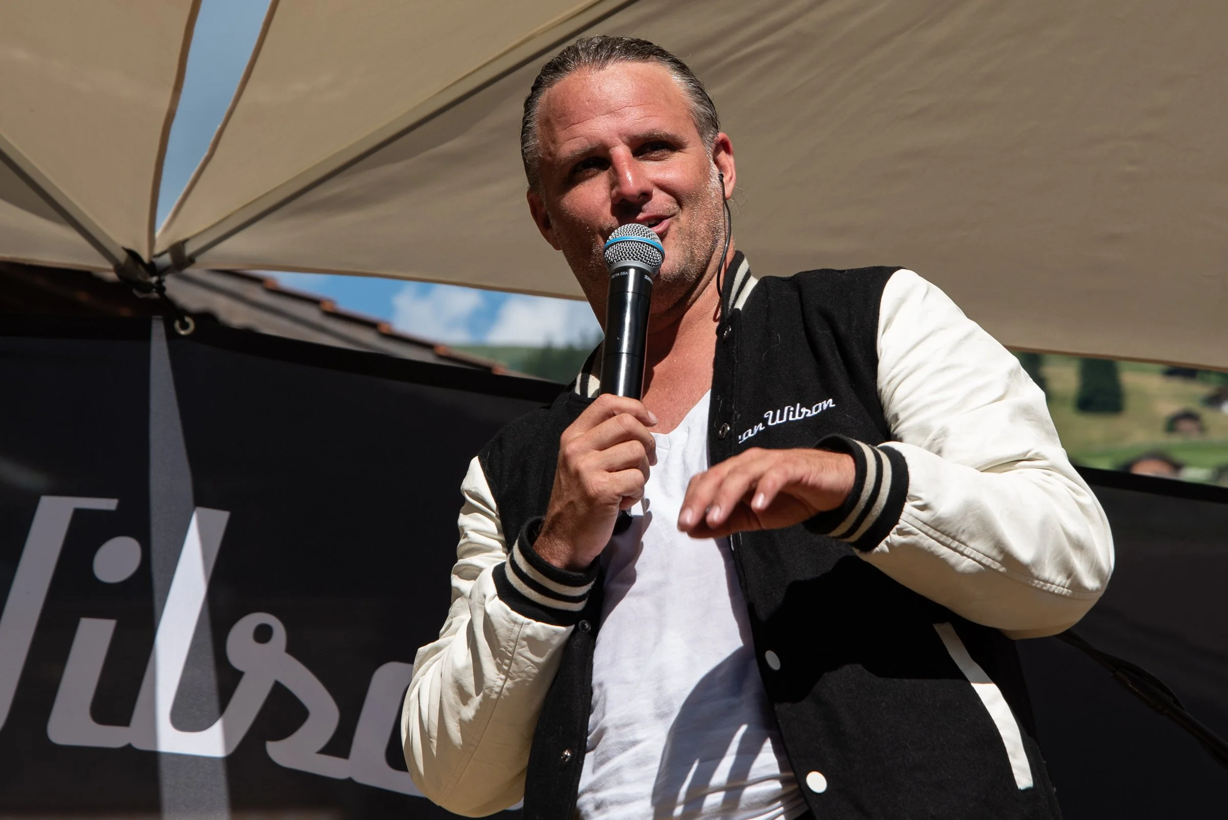 Ein Mann hält ein Mikrofon in der Hand und spricht, während er im Freien steht, unter einem Sonnenschirm, mit einer ländlichen Landschaft im Hintergrund.