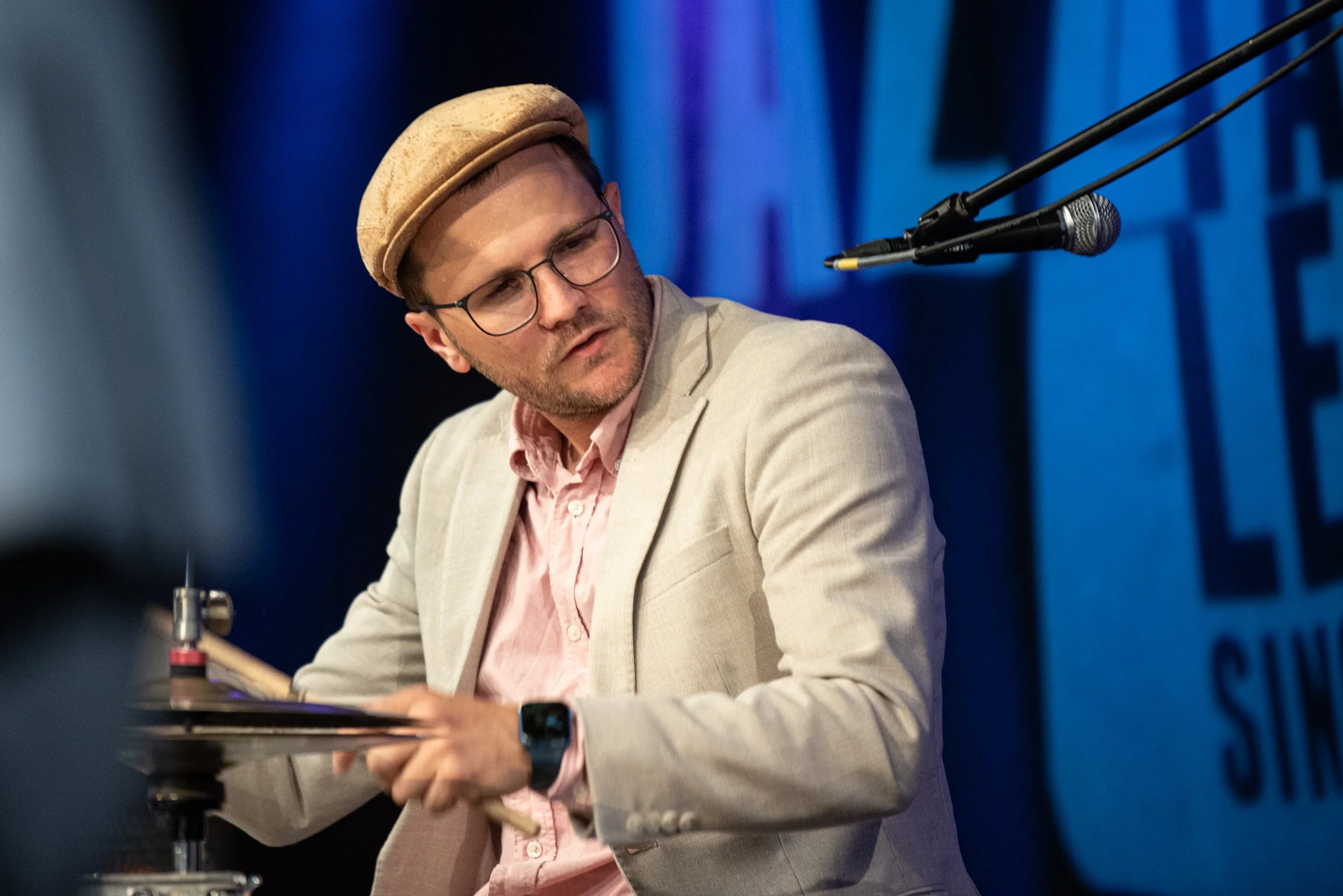 Mann spielt Schlagzeug bei einem Konzert, trägt Brille, Hut und Anzug, auf der Bühne mit dunklem Hintergrund.