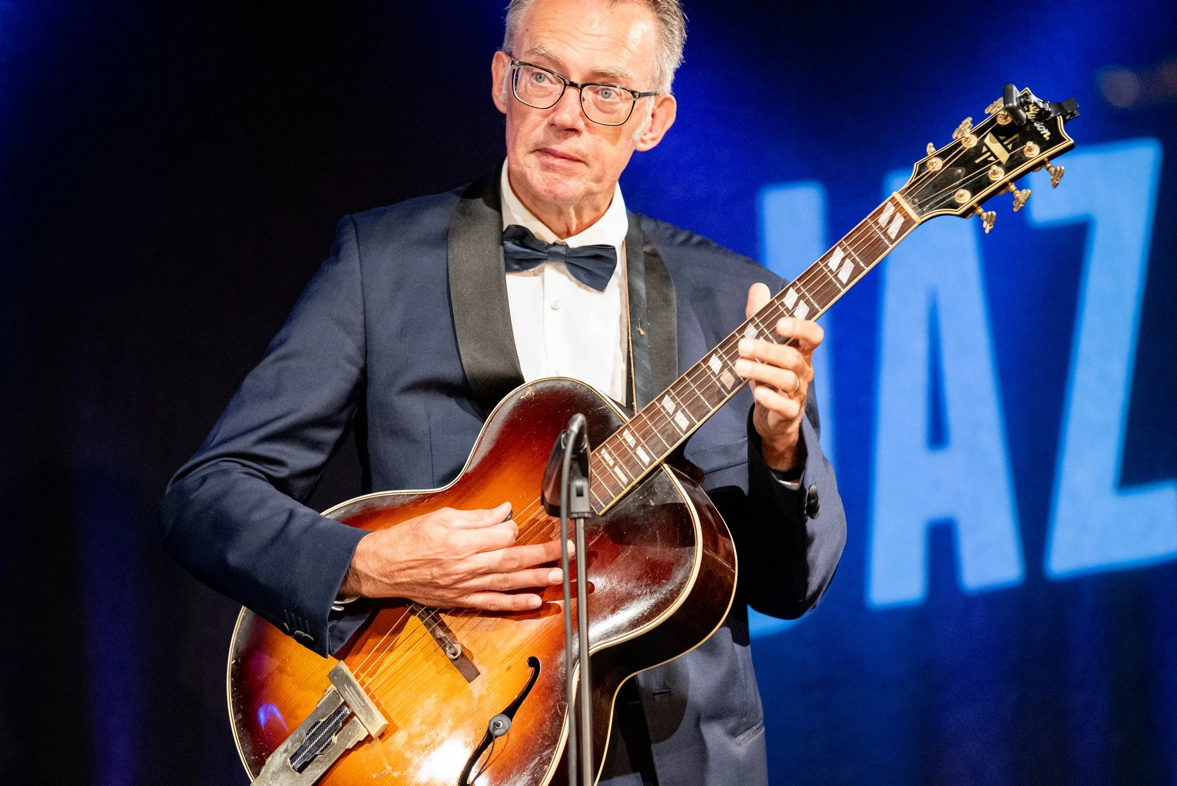 Ein älterer Mann in einem smoking trägt eine Brille und spielt Gitarre auf einer Bühne mit einem blauen Hintergrund, auf dem in großen Buchstaben 'KAZ' zu sehen ist.