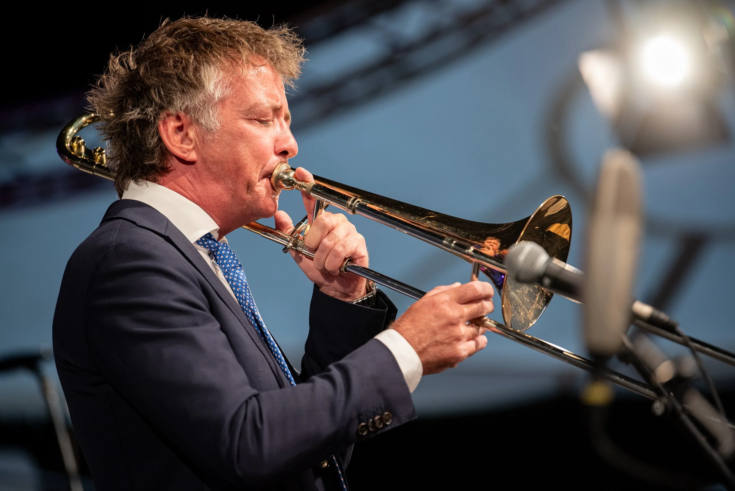 Ein Mann spielt auf einem goldenen Trompeteninstrument bei einem Auftritt, trägt einen dunklen Anzug und eine blaue Krawatte, vor einem Mikrofon.