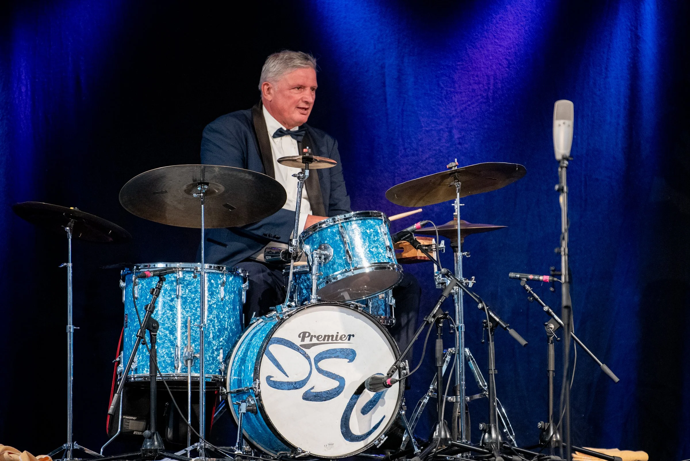 Ein älterer Mann in formeller Kleidung spielt auf einem blauen Schlagzeug auf einer Bühne mit dunklem, bläulichem Hintergrund.