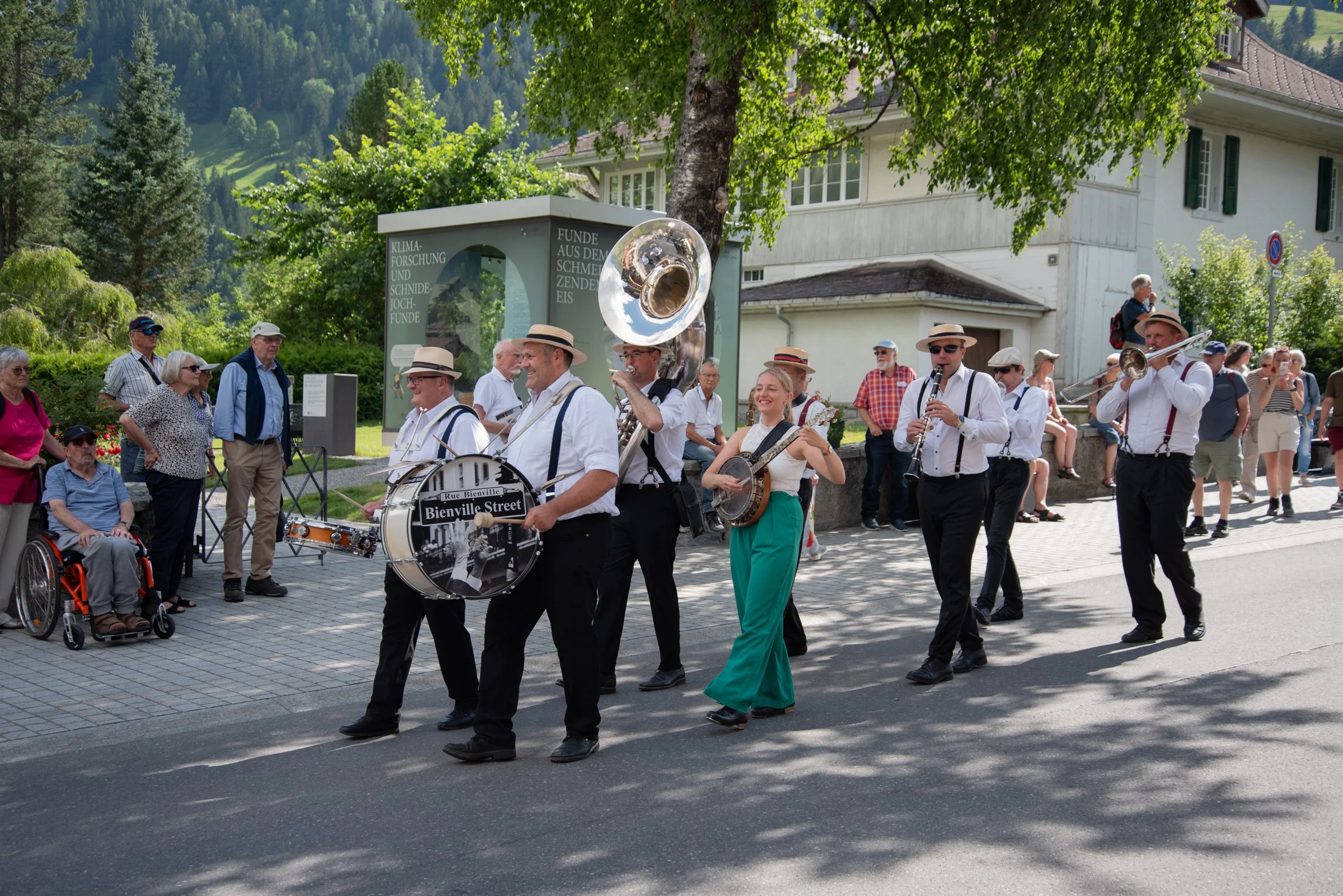 Eine Gruppe von Musikern in weißen Hemden und schwarzen Hosen, die eine Straßenparade in einem Grünen und sonnigen Gebiet mit Bäumen und Bergen im Hintergrund spielen, während Menschen zuschauen.