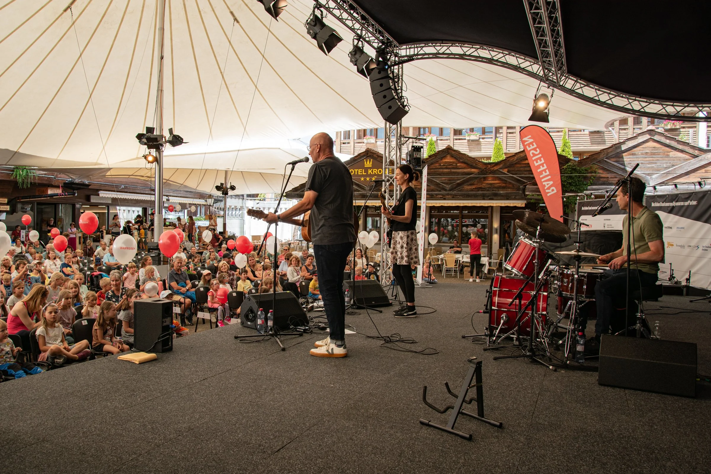 Musiker auf einer Bühne, die unter einem großen Zelt im Freien spielt, vor einer Menschenmenge bei einem Fest mit roten und weißen Ballons.